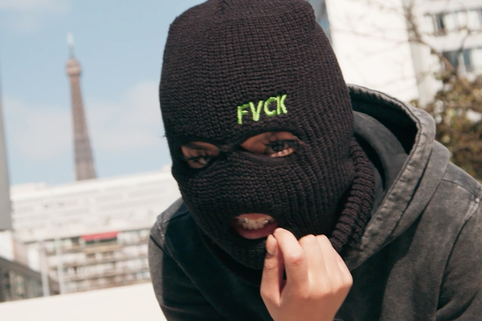 Une personne portant un masque de ski noir avec le mot 'FUCK' en vert brodé, en tenue décontractée, dans une ville avec la Tour Eiffel en arrière-plan.