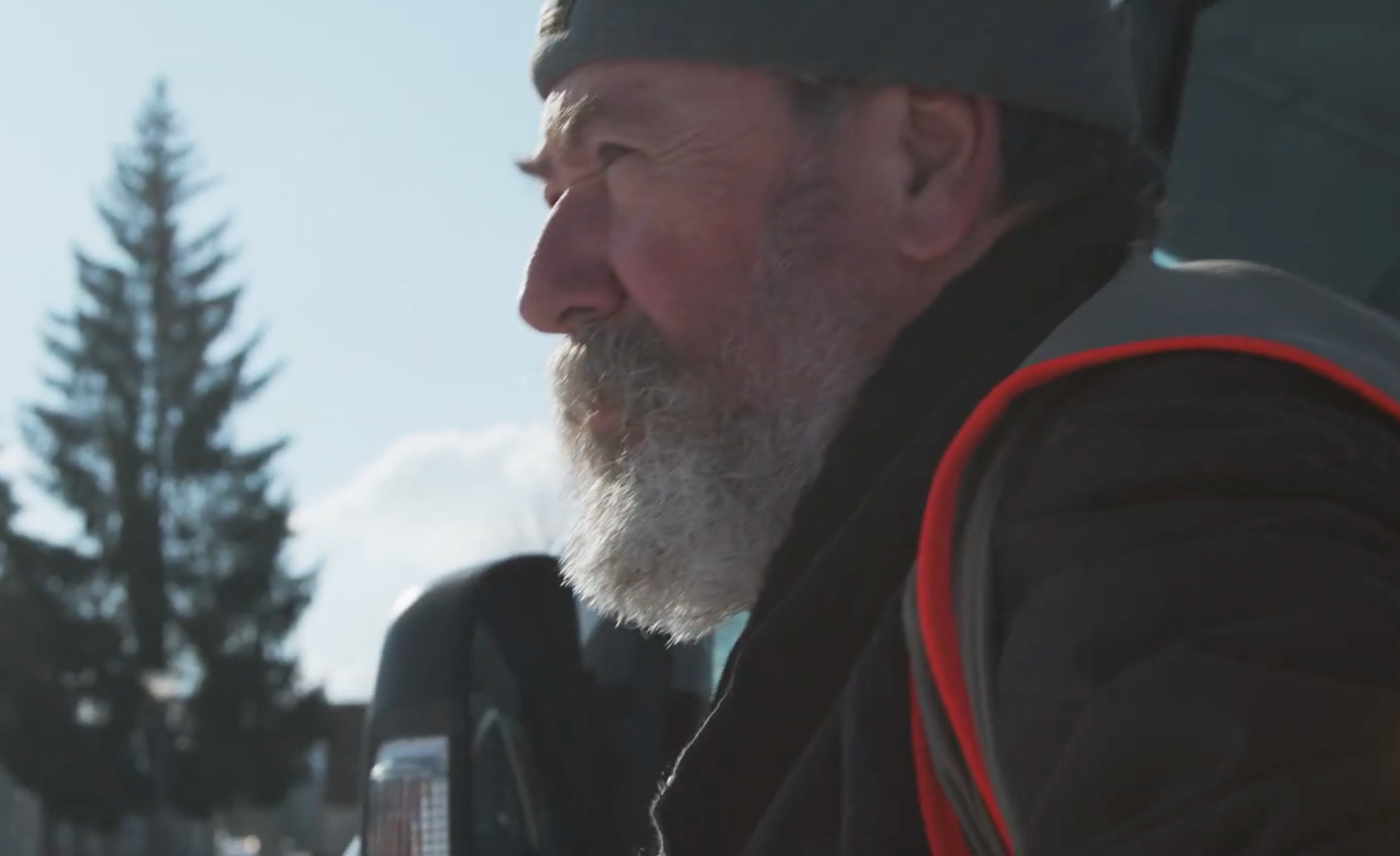 Un homme âgé avec une barbe grise, portant un bonnet et une veste noire avec une poche rouge, se trouve à l'extérieur avec un arbre en arrière-plan et un ciel ensoleillé.