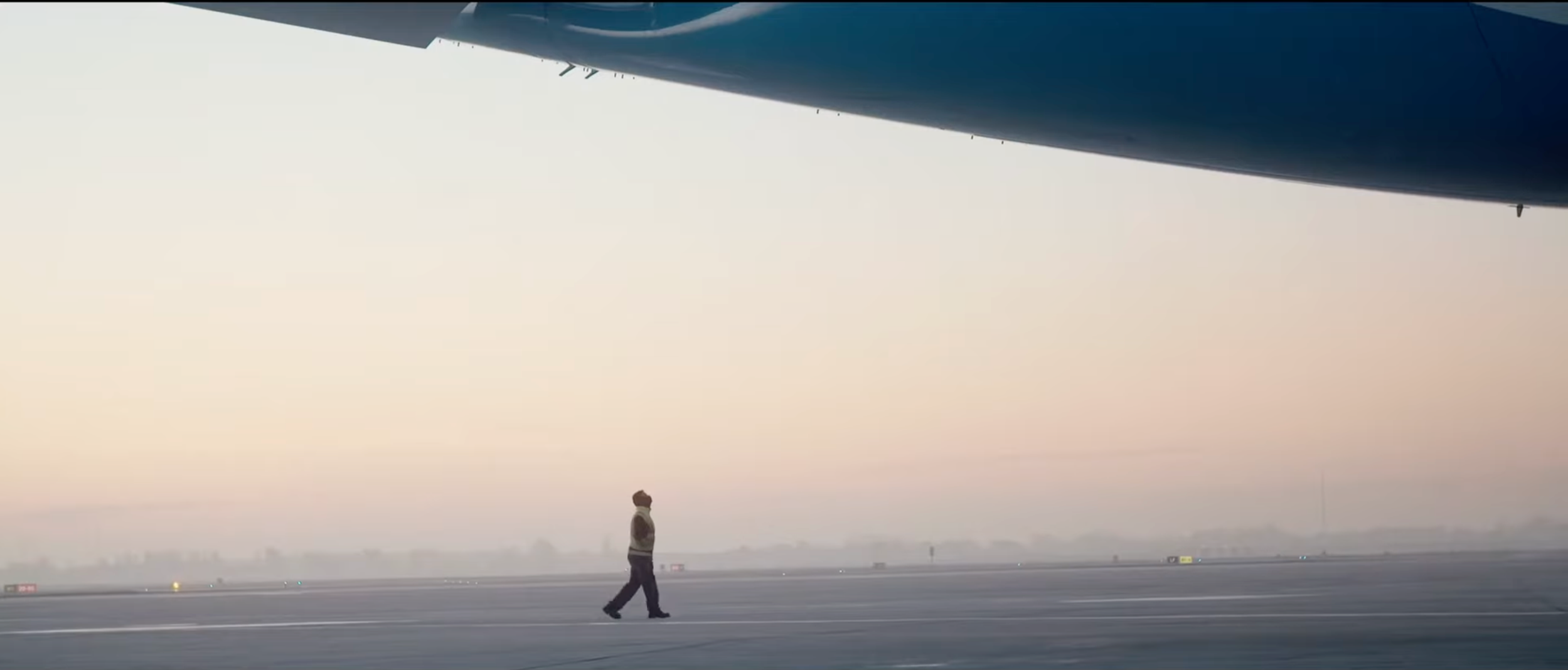 Un homme marche sur un tarmac d'aéroport avec un avion stationné au-dessus de lui, dans un ciel nuageux au crépuscule.