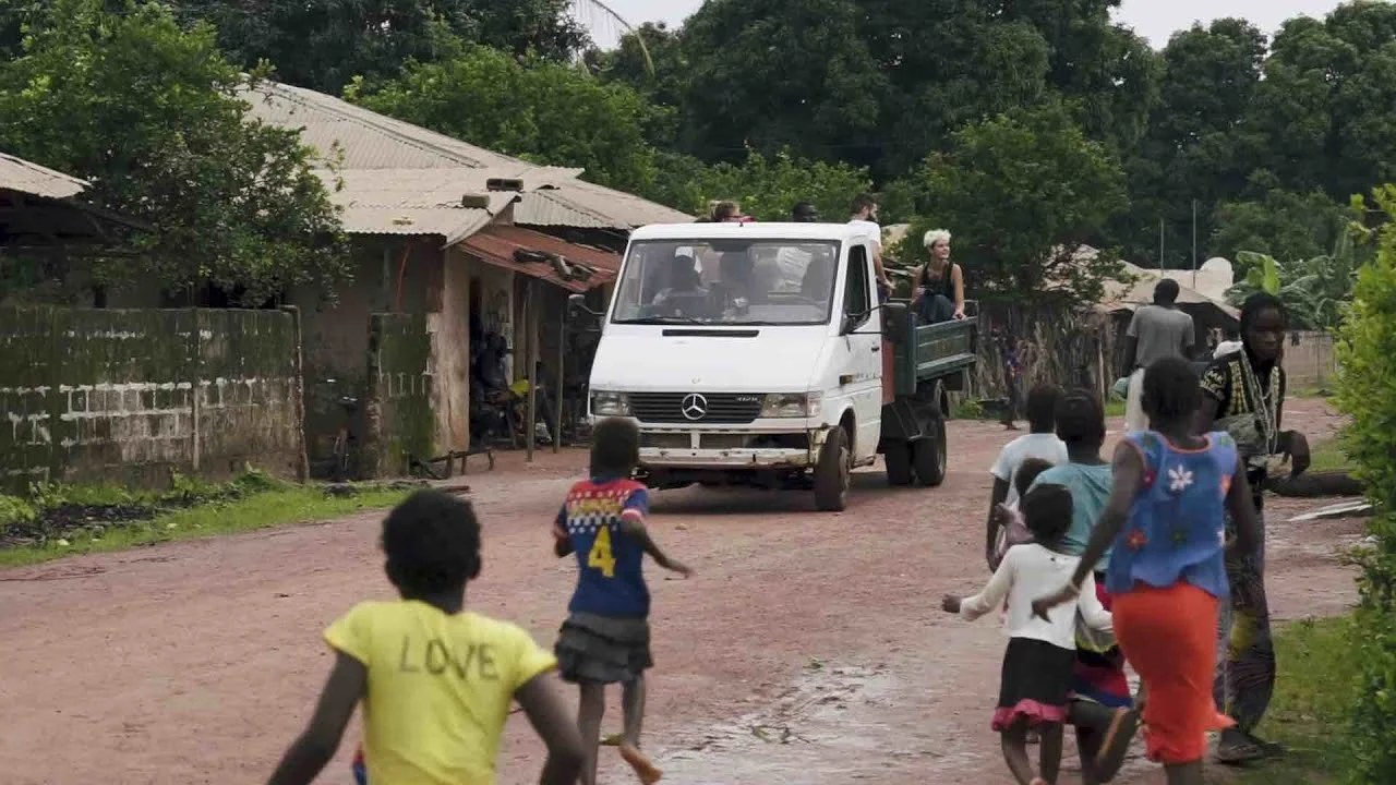 Enfants jouant dans une rue poussiéreuse avec une grande camionnette blanche Mercedes en mouvement, dans un environnement rural avec des huttes en toiture de tôle et de la végétation dense en arrière-plan.