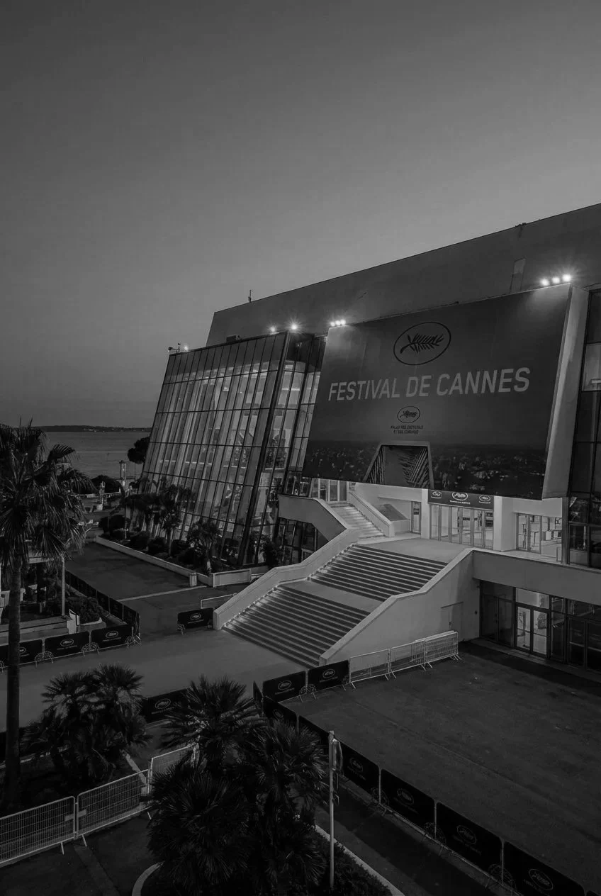 Bâtiment moderne du Festival de Cannes avec un grand panneau publicitaire et des escaliers, entouré de palmiers, près de la mer, en noir et blanc.