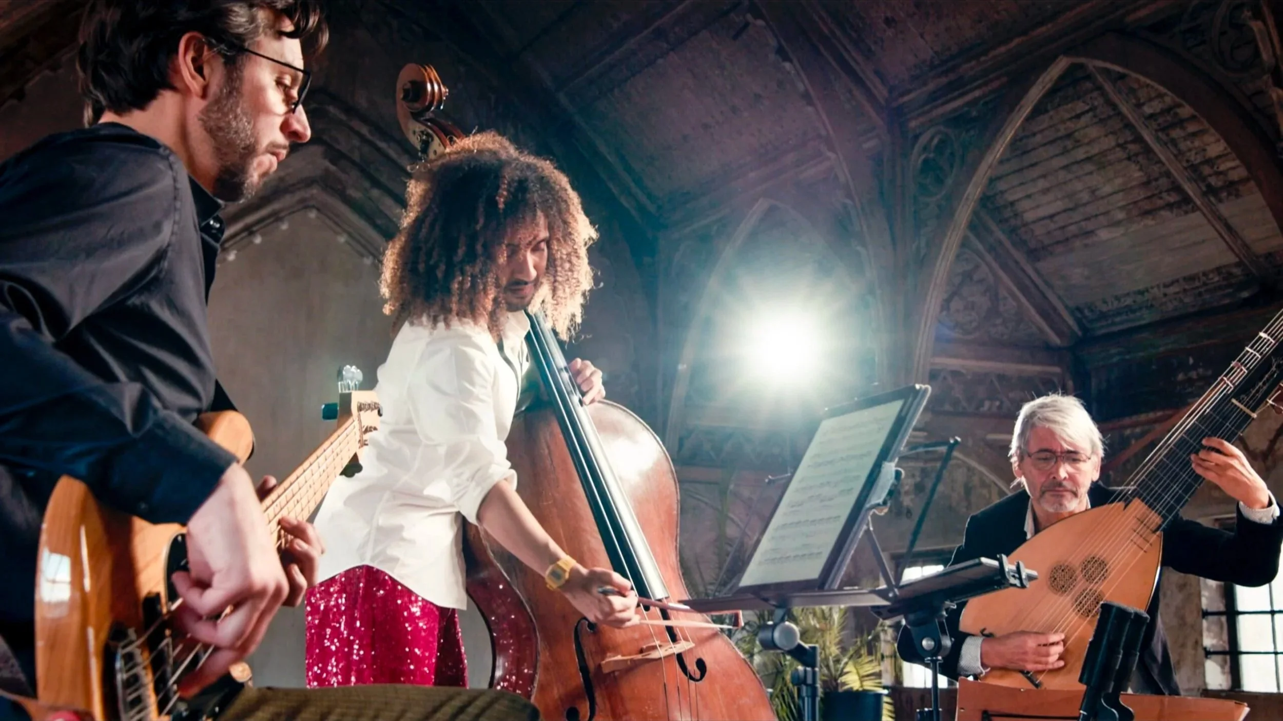 Vier muzikanten spelen samen in een concertzaal met houten plafonds en grote ramen, gebruikmakend van gitaar, cello en andere instrumenten.