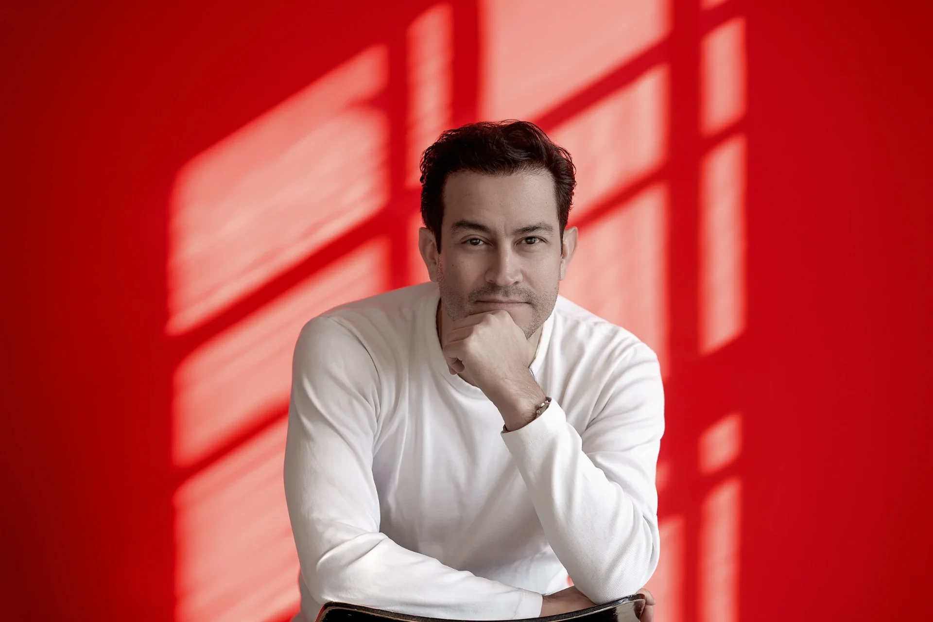 Man met korte donkere haar, witte trui, poseert met hand onder kin voor een rode achtergrond met schaduwen van raamkozijnen.