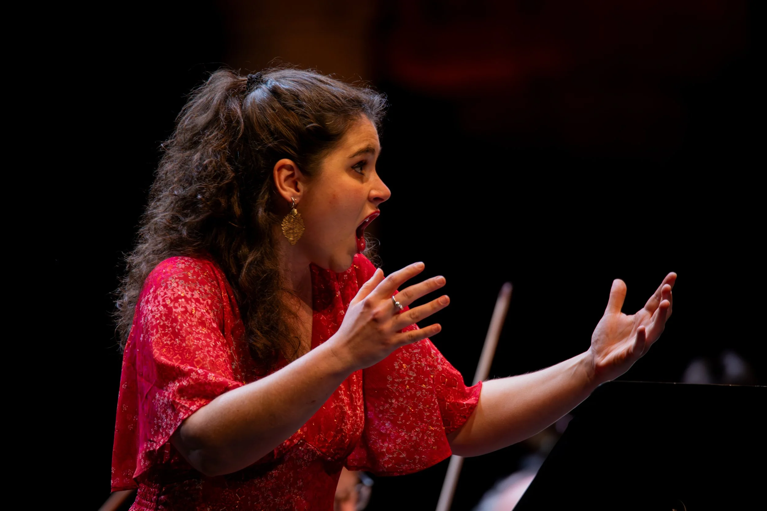 Vrouw met lang, krullend haar en grote gouden oorbellen, gekleed in een rode, glanzende jurk, zingt of spreekt enthousiast met haar mond open en handen gestrekt.