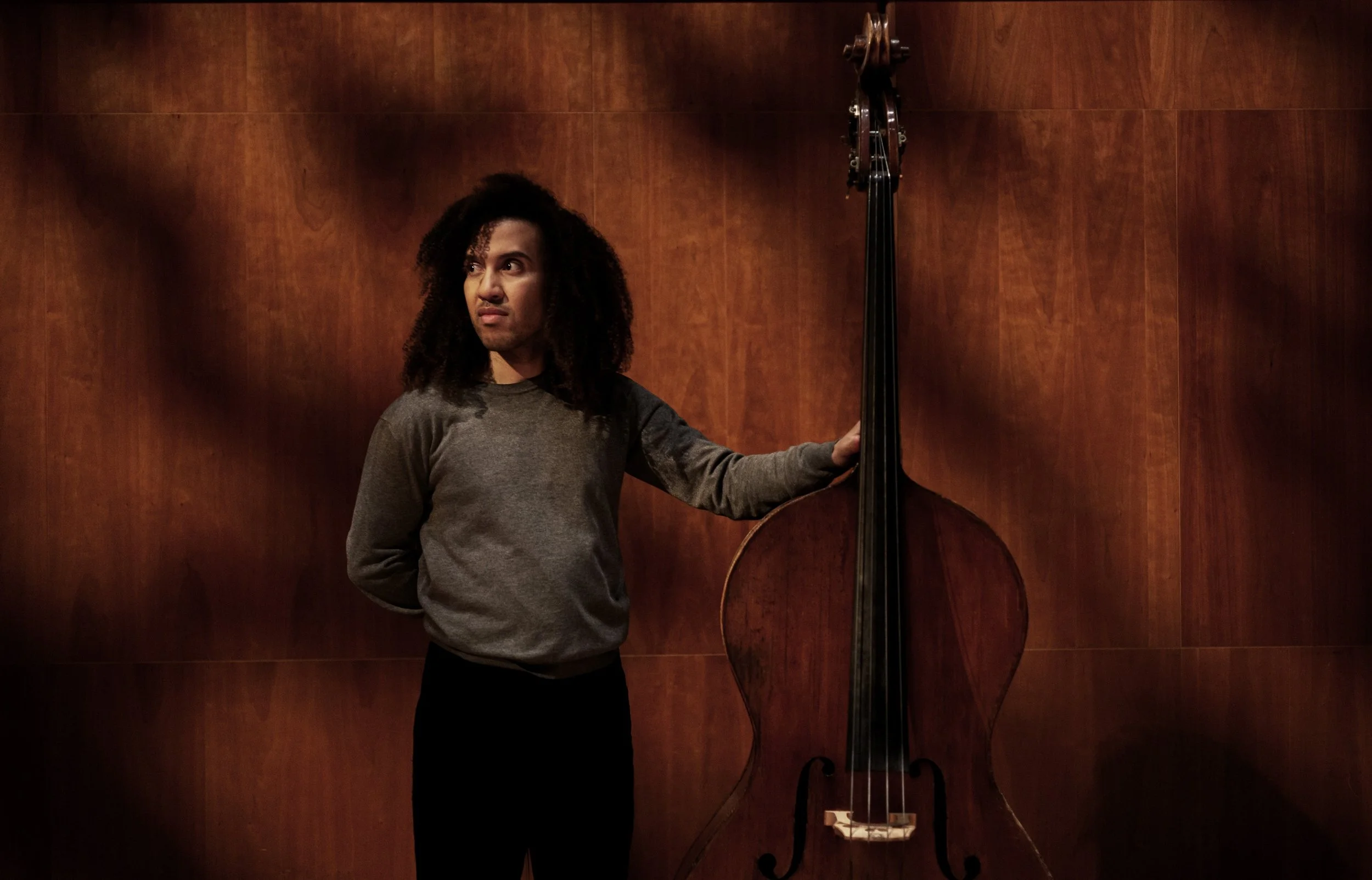 Een persoon met curly haar staat naast een grote contrabas, met een houten paneel op de achtergrond.