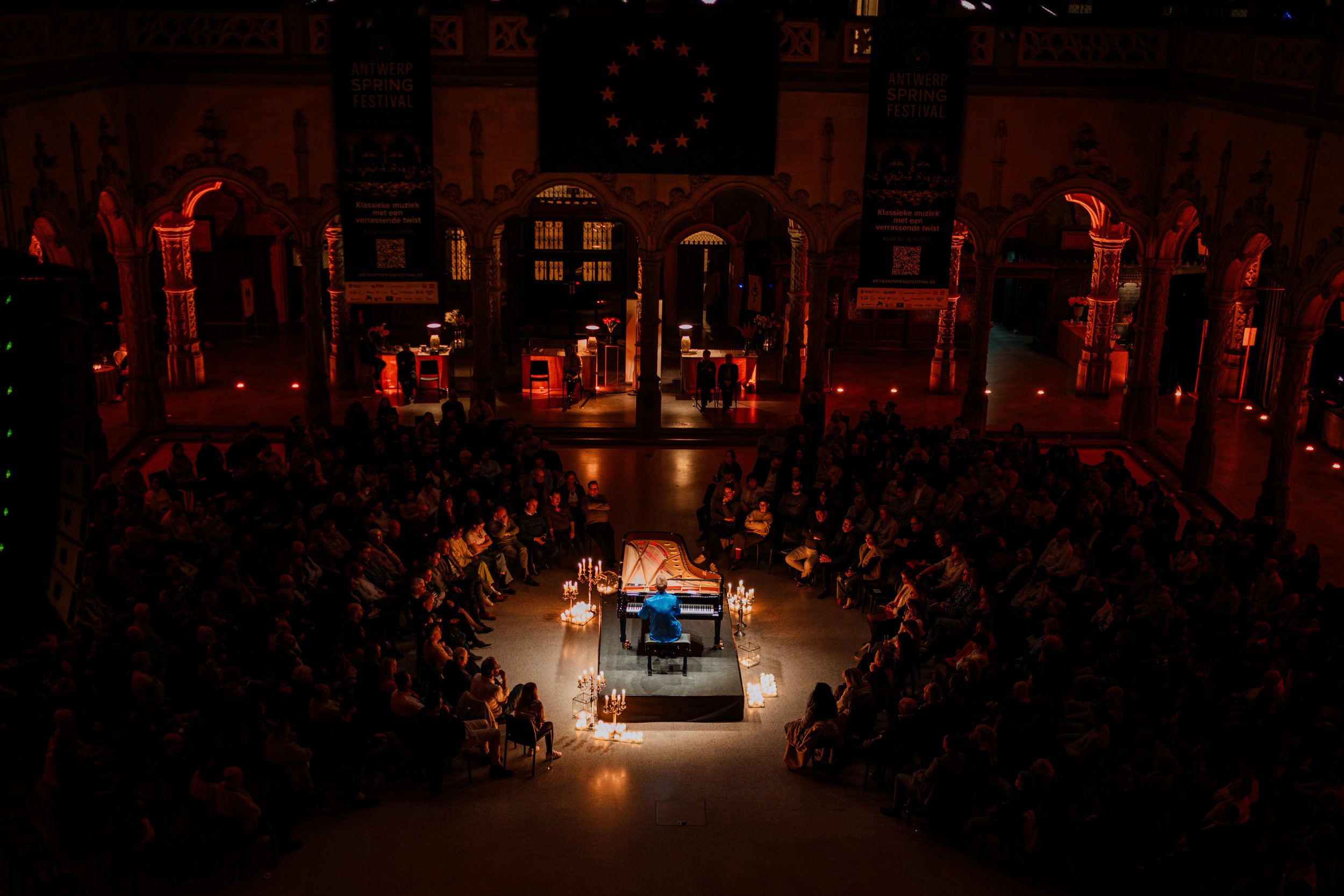 Een concert in een grote zaal met een pianist die op een zwarte piano speelt, omringd door candelaren met brandende kaarsen, met een publiek dat kijkt. In de achtergrond hangt een tekstbord en een vlag met sterren en cirkelvormige sterren. De zaal heeft een middeleeuwse architectuur met ornamenten en bogen.