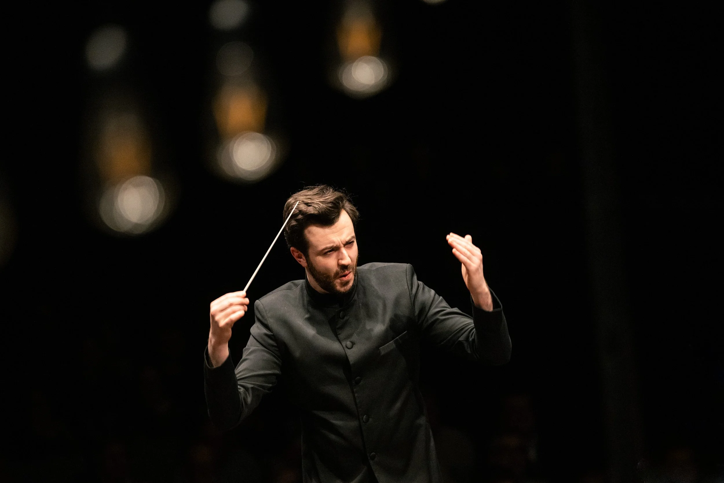 Dirigent die orkestermuziek dirigeert, met een concentratieve uitdrukking, tegen een zwarte achtergrond met onscherpe lichteffecten.