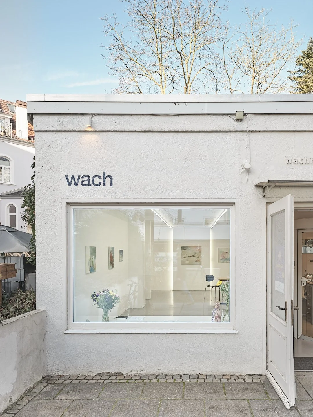 Außenansicht der Bremer Galerie ‚wach Kunstraum‘ mit großem Fenster, Kunstwerken und offenem Eingang; exterior view of the Bremen gallery ‘wach Kunstraum’ with a large window displaying artworks and an open door.