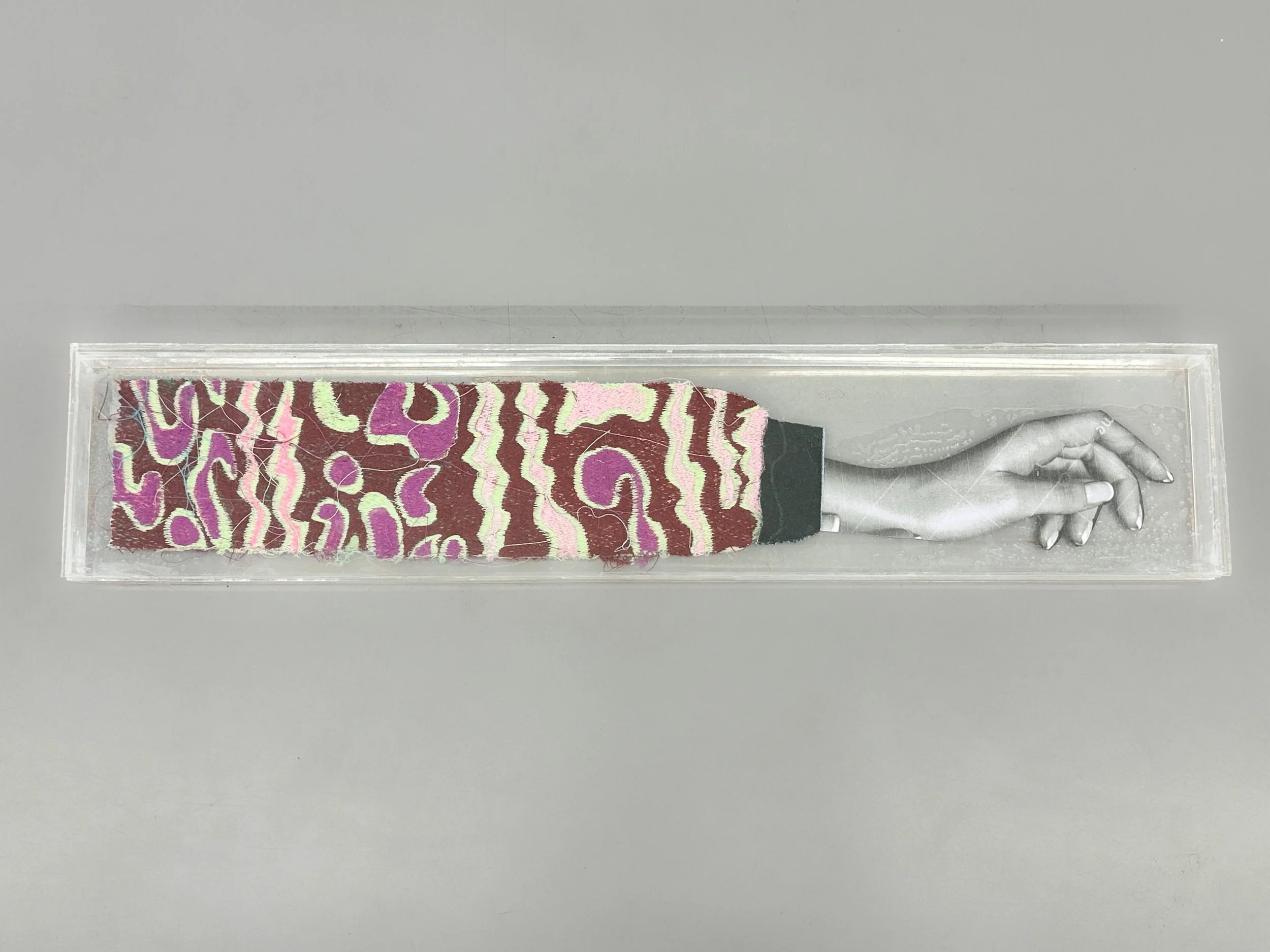 Handmade sculpture of red camouflage textiles attached to a black-and-white photocopied hand gesture, enclosed in a plexiglass box.