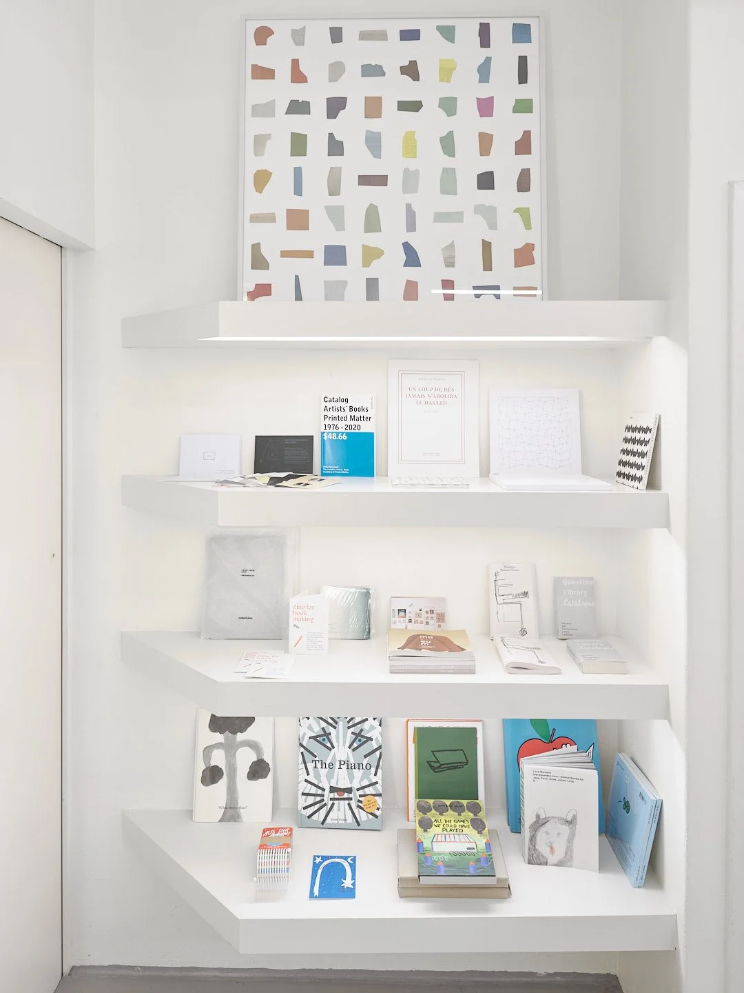Publikationsbereich mit weißen Regalen voller Kunstbücher, Kinderbücher und Magazine nahe einer Tür; artist publication area with white shelves holding art books, children’s books, and magazines near a doorway.