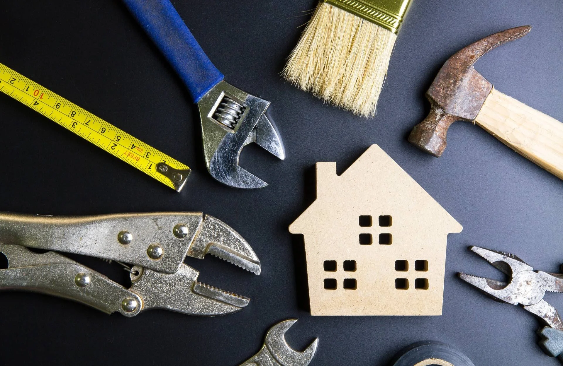 A spreadsheet tape measure, adjustable wrench, paintbrush, hammer, pliers, wrench, a small wooden house model, and some tape on a black surface.