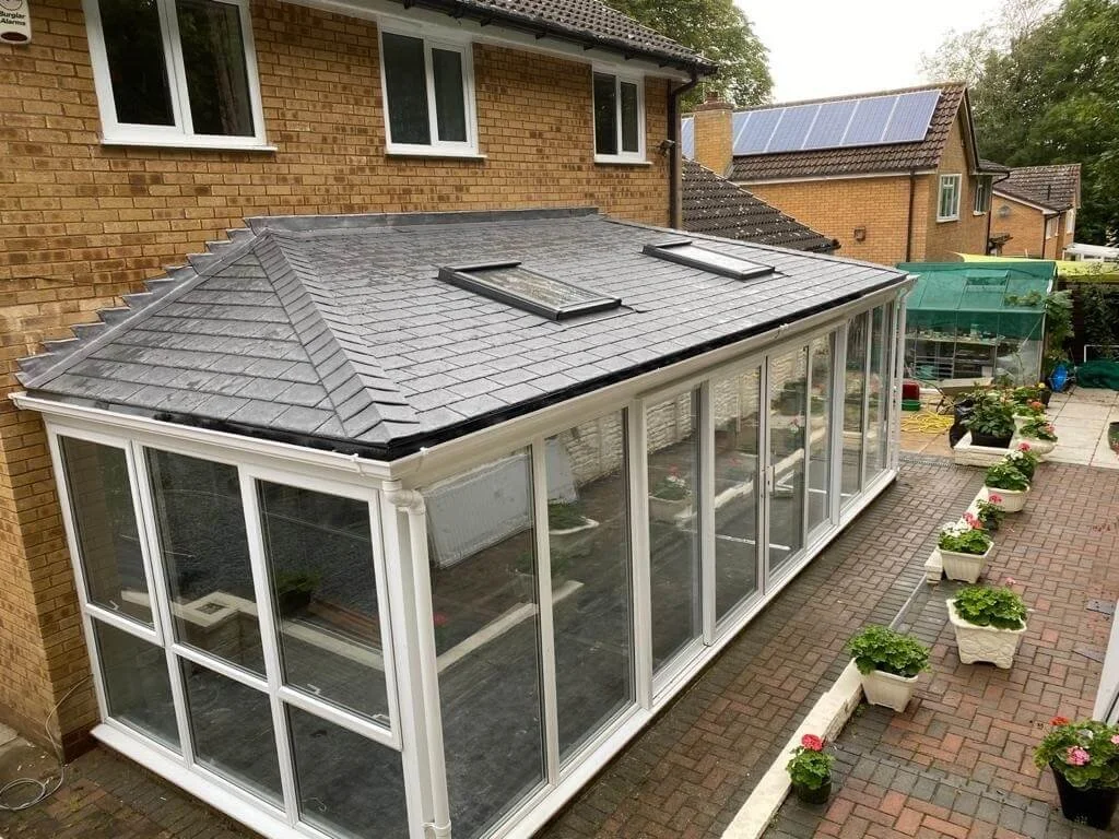 A backyard with a brick house featuring a conservatory or sunroom with large glass windows. The roof of the conservatory has two skylights and is covered with gray shingles. Potted plants line the brick pathway alongside the conservatory, and there are additional houses with solar panels in the background.