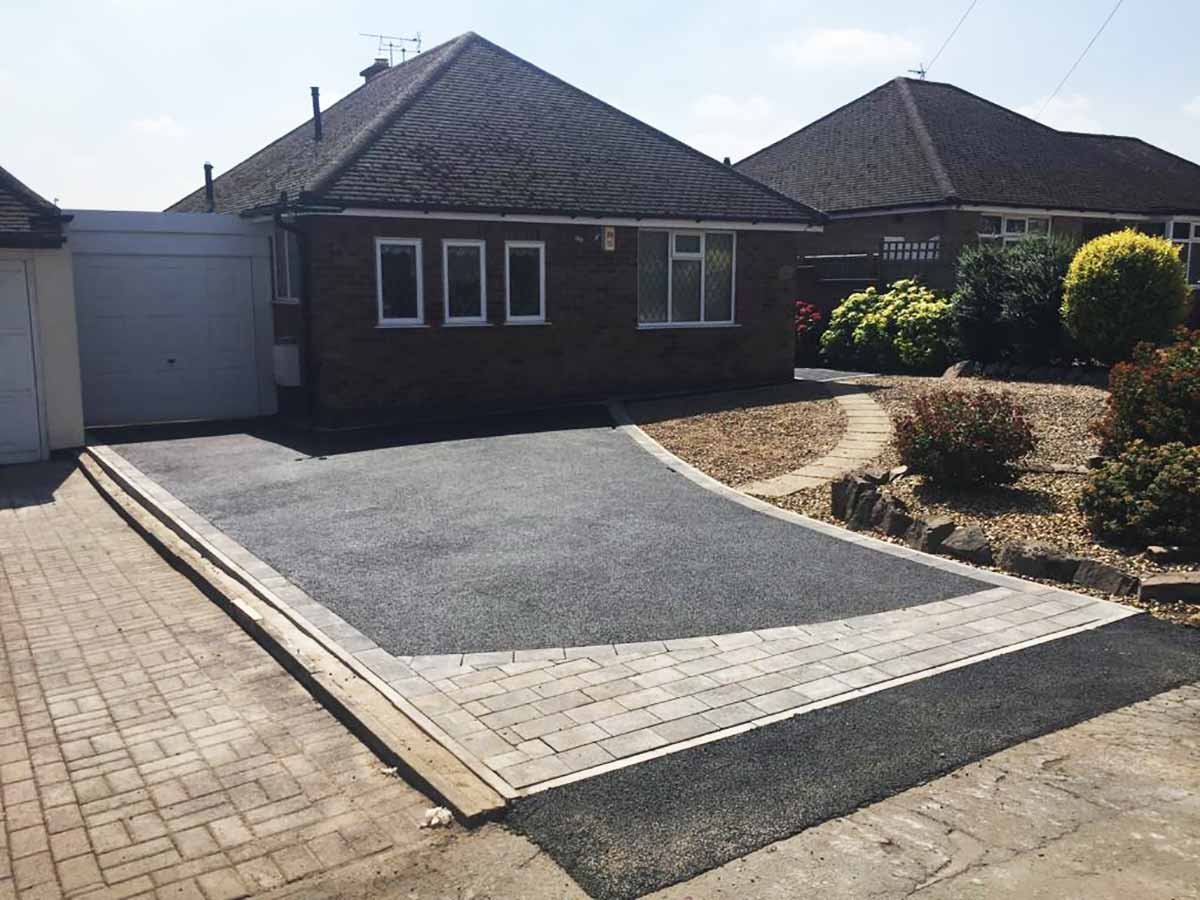 A residential driveway with a newly paved section, bordered by brick pavers, leading to a garage, with a small walkway and landscaped garden with bushes and flowers to the right.