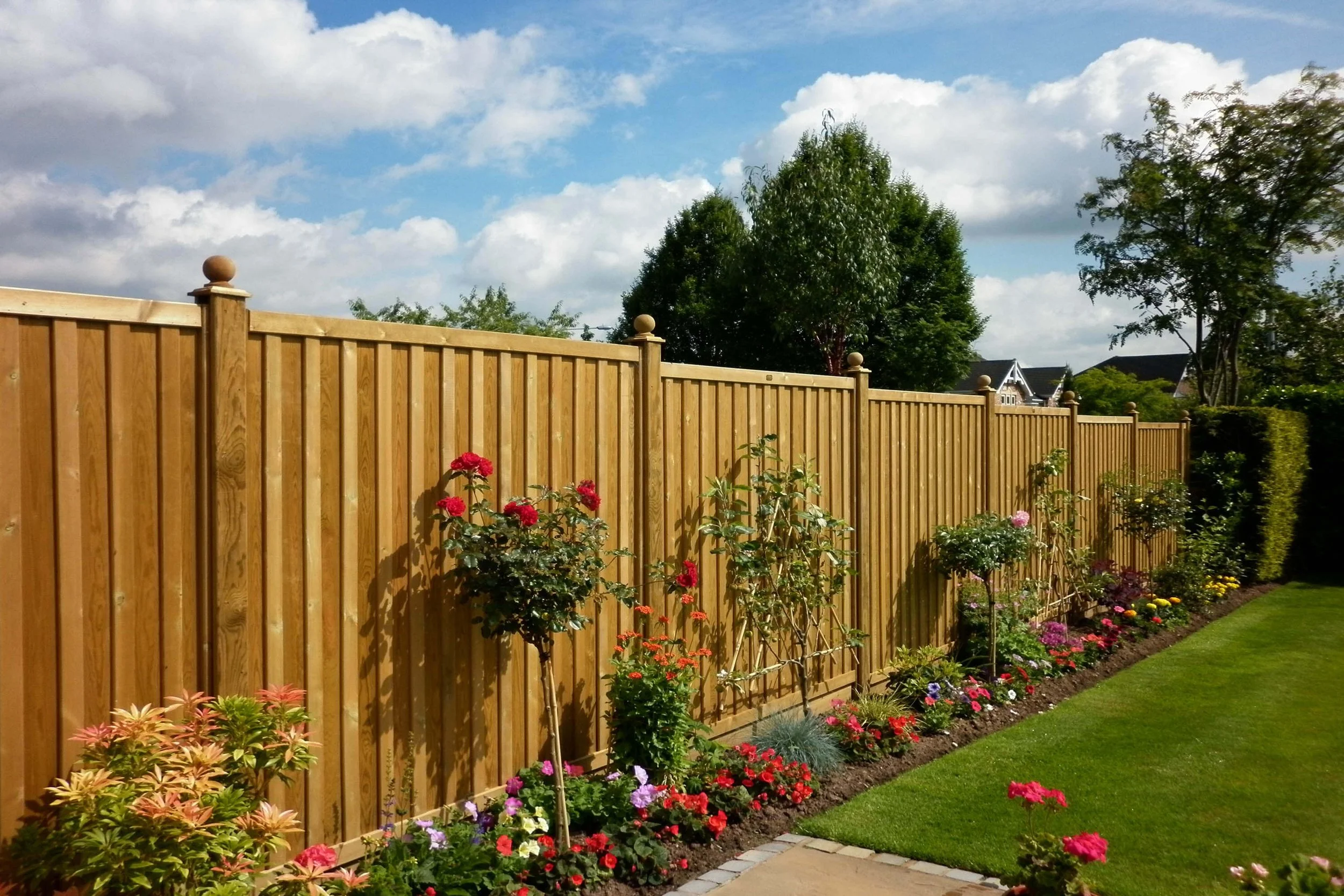 A wooden garden fence with a flower bed featuring colorful blooming flowers and shrubs, beside a well-manicured grassy area under a partly cloudy sky.
