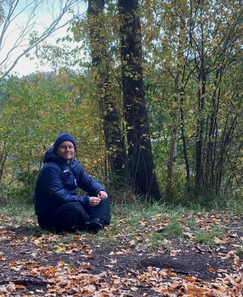 Lukas sitting on the ground in a wooded area with autumn leaves, wearing a dark jacket and beanie, smiling at the camera.