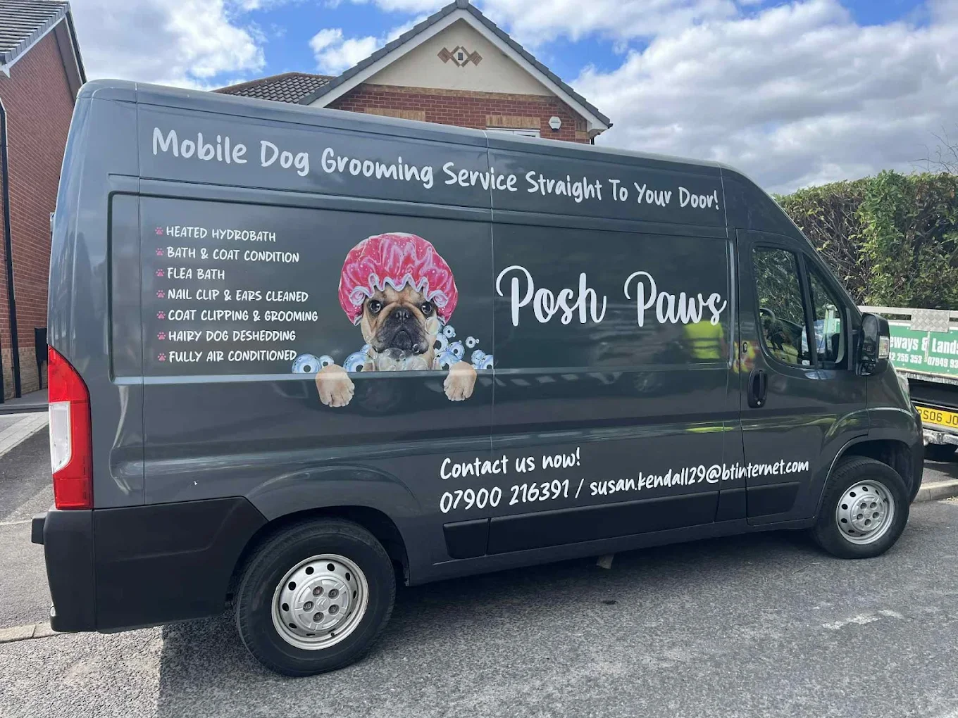 A dark grey van for a mobile dog grooming service called 'Posh Paws' parked on a street with a house in the background. The van features a graphic of a dog wearing a pink shower cap, with the company logo and contact information on the side.