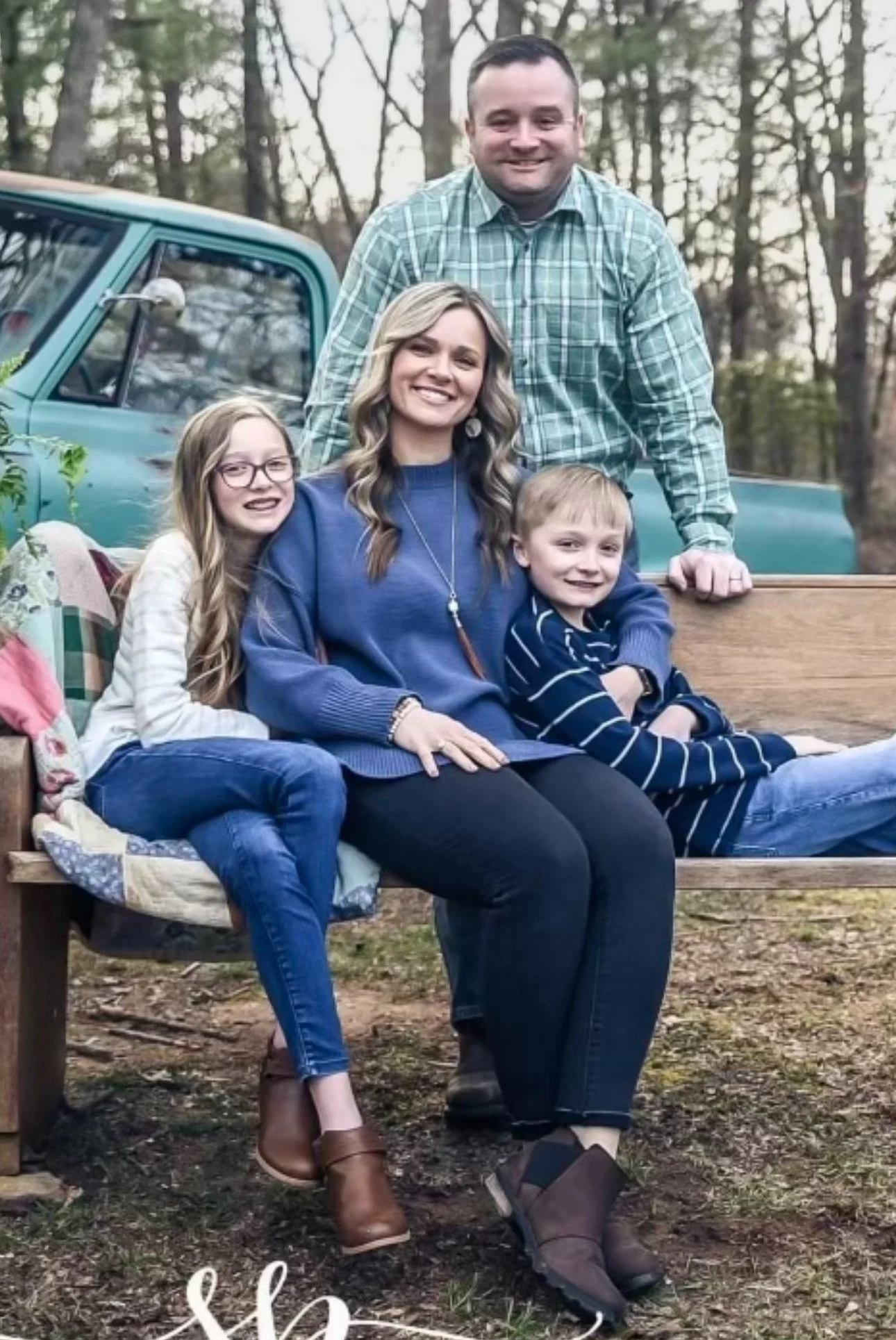 A family of four sitting and standing on a wooden bench in a forest outdoors, smiling at the camera. The family consists of a man, woman, and two children.