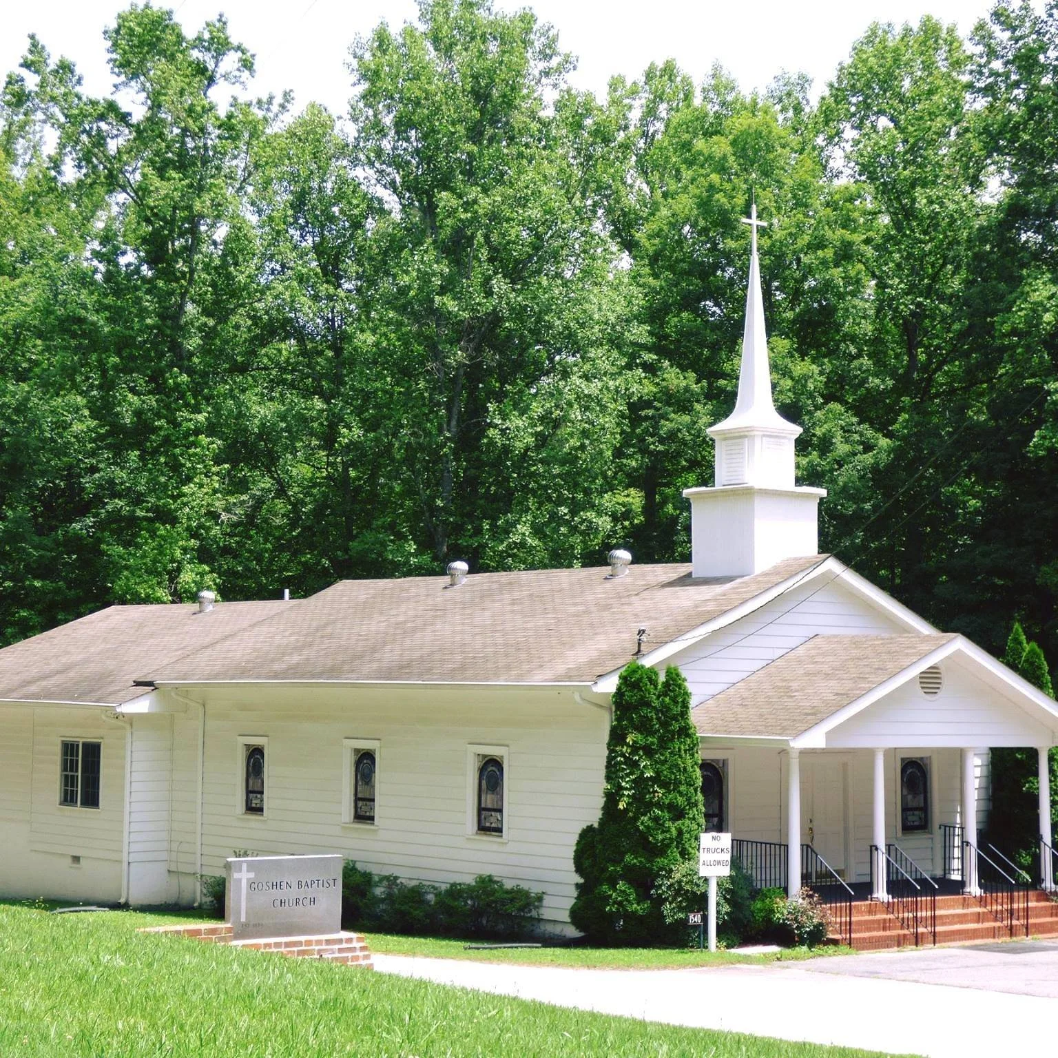 A small white church with a steeple and cross, surrounded by green trees, with steps leading to the entrance and a sign reading 'Goshen Baptist Church' in the front yard.