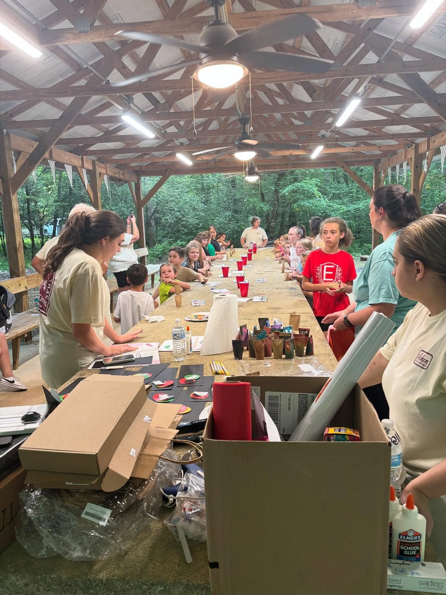 A group of children and adults gathered around a long table inside a wooden pavilion outdoors, with some people standing and others sitting, engaged in activities. The pavilion has a high roof with ceiling fans and bright lights, and the background shows green trees.