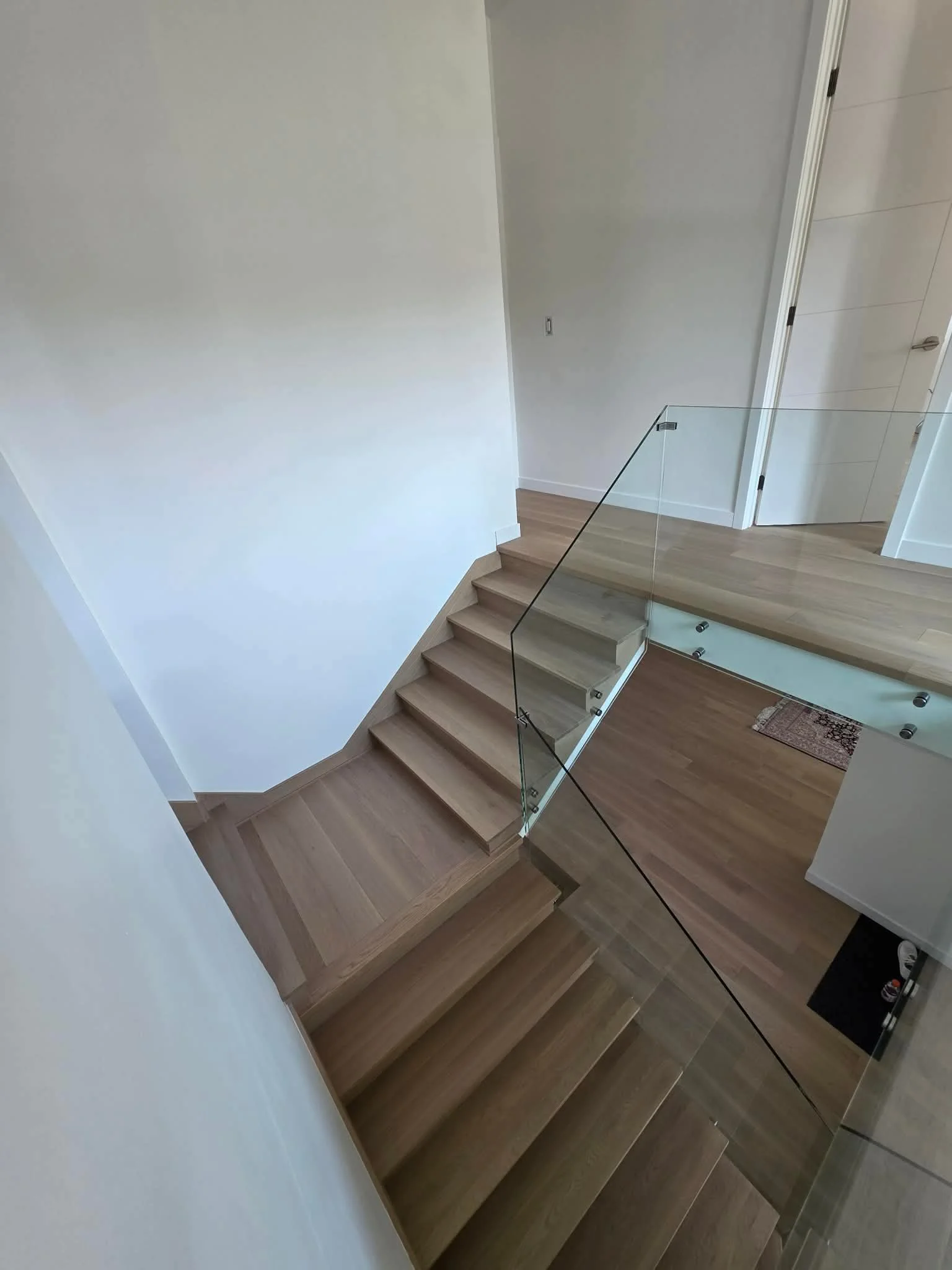 Interior view of a house staircase with wooden steps, glass railing, and a beige wall at the top of the stairs.
