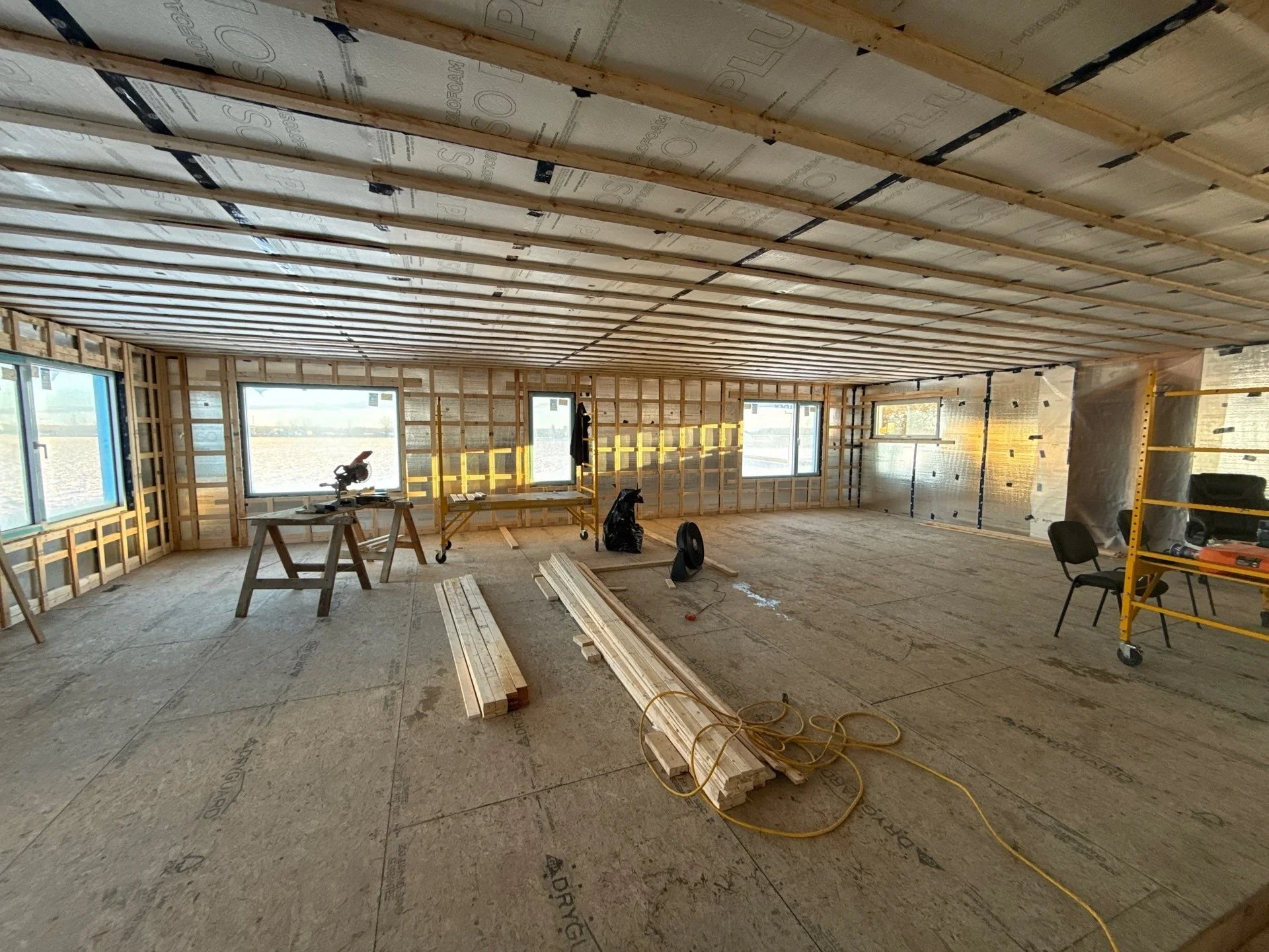 Interior of a house under construction with exposed roof framing, window openings, unfinished walls, construction tools, and materials on the floor.