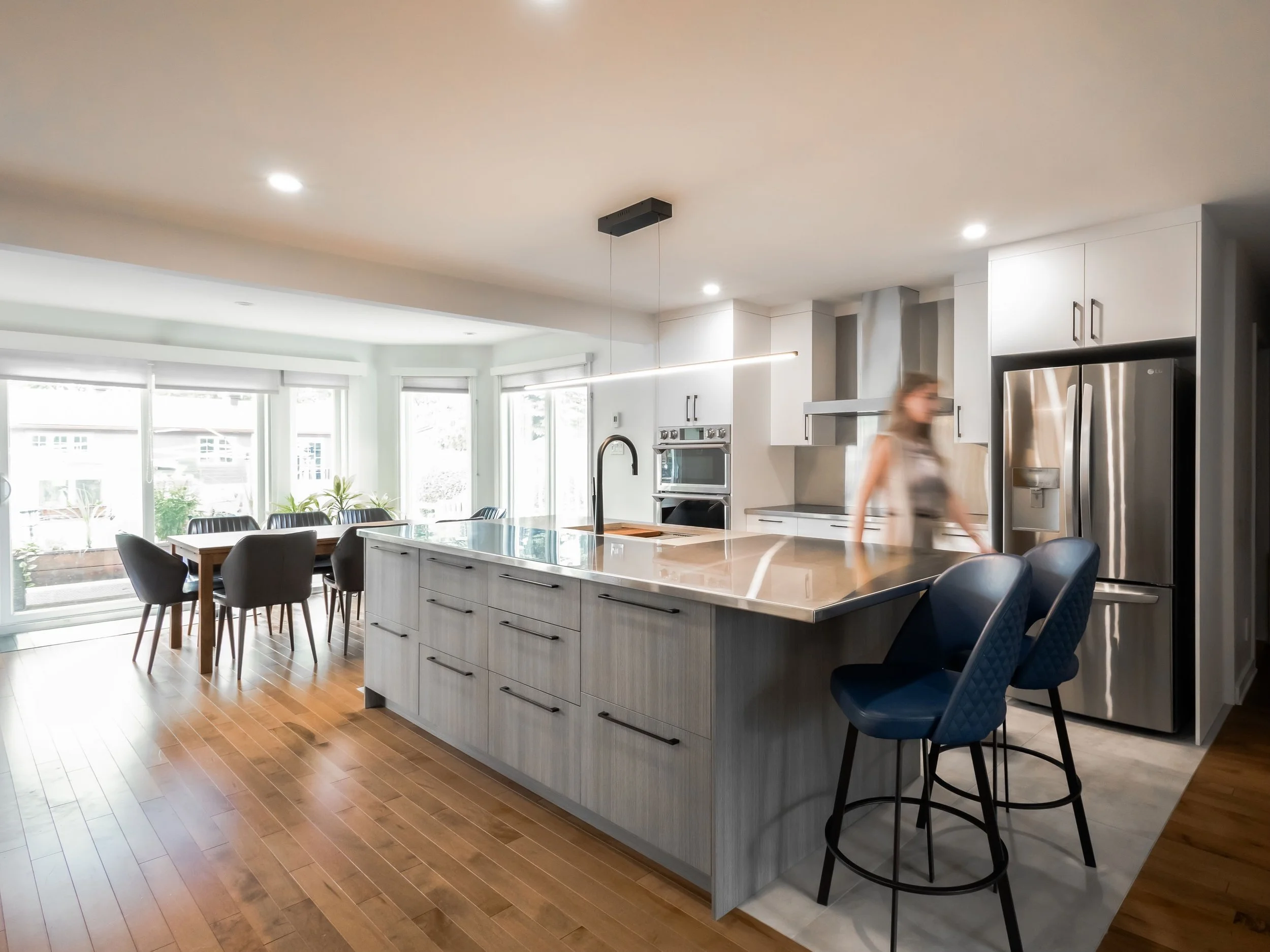 Bright modern kitchen with white cabinetry, stainless steel appliances, a large island with a marble countertop, and a dining area with a table and black chairs near large windows.