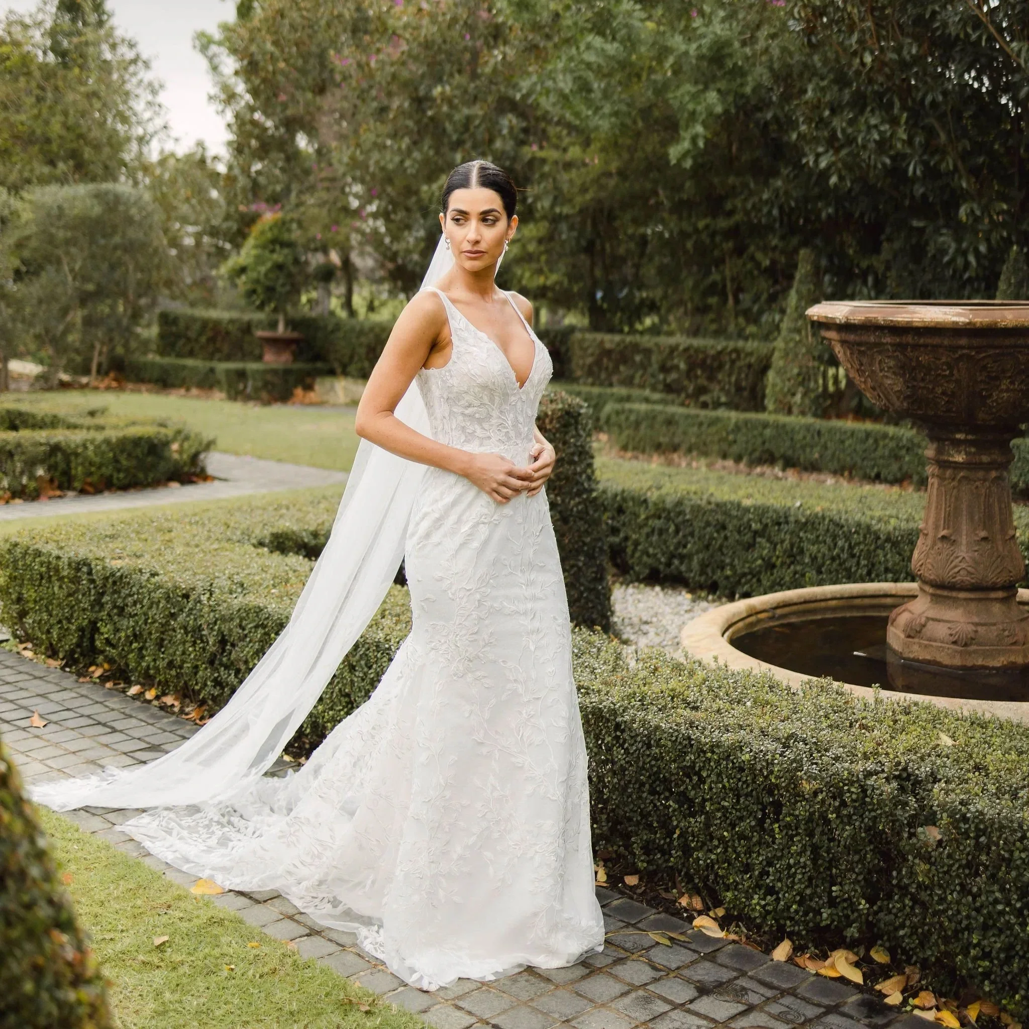 Bride modeling a boho lace wedding gown with a deep V neckline and long veil in a romantic garden setting, showcasing designer bridal styles available at Ivory Aisle Bridal.
