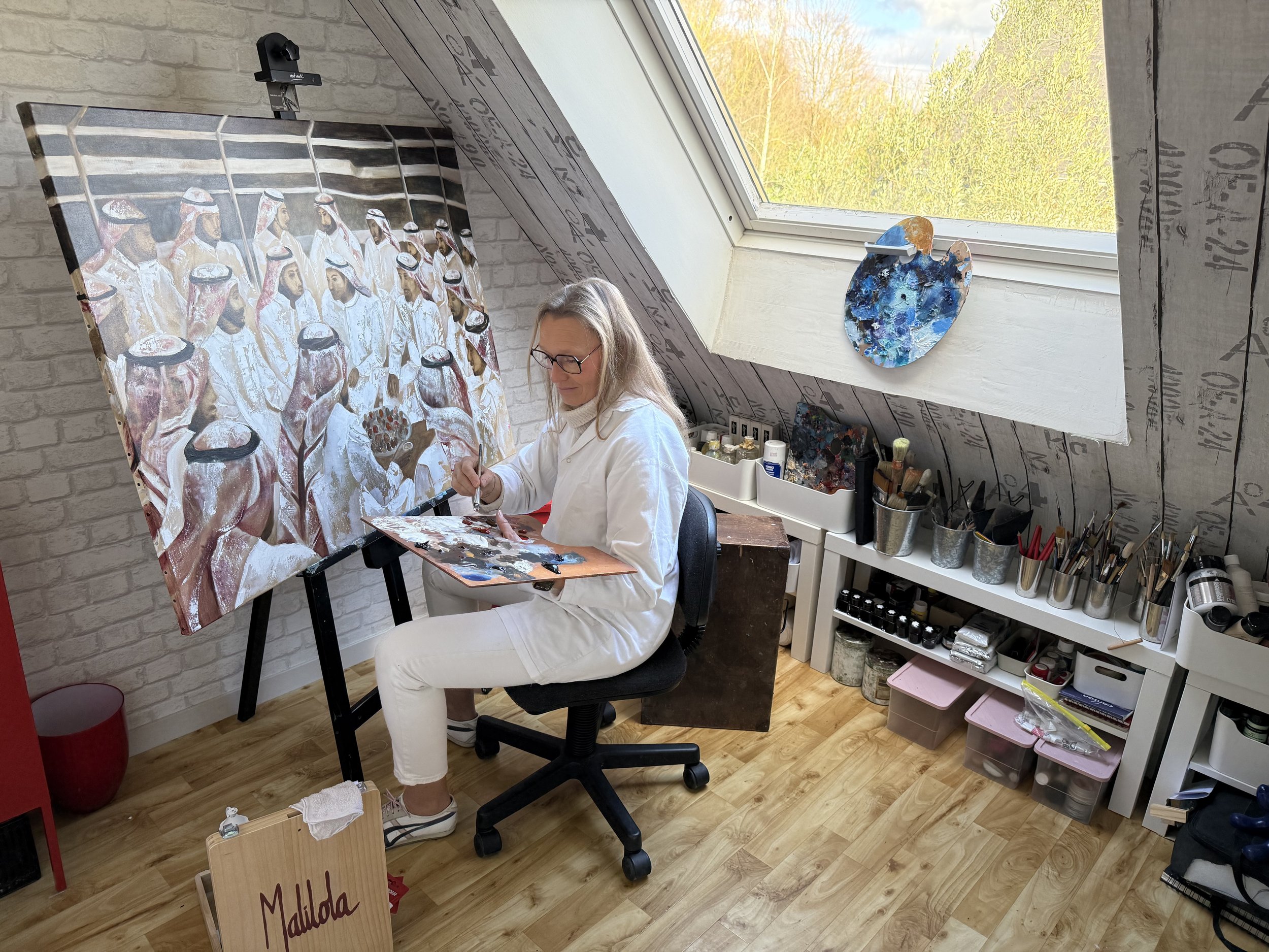 Marie-Laure HUBERT, Malilola, the artist is painting a large canvas in a bright attic studio, depicting a scene in a majlis, Saudi traditional gathering place, with many Saudi men wearing white traditional garments.