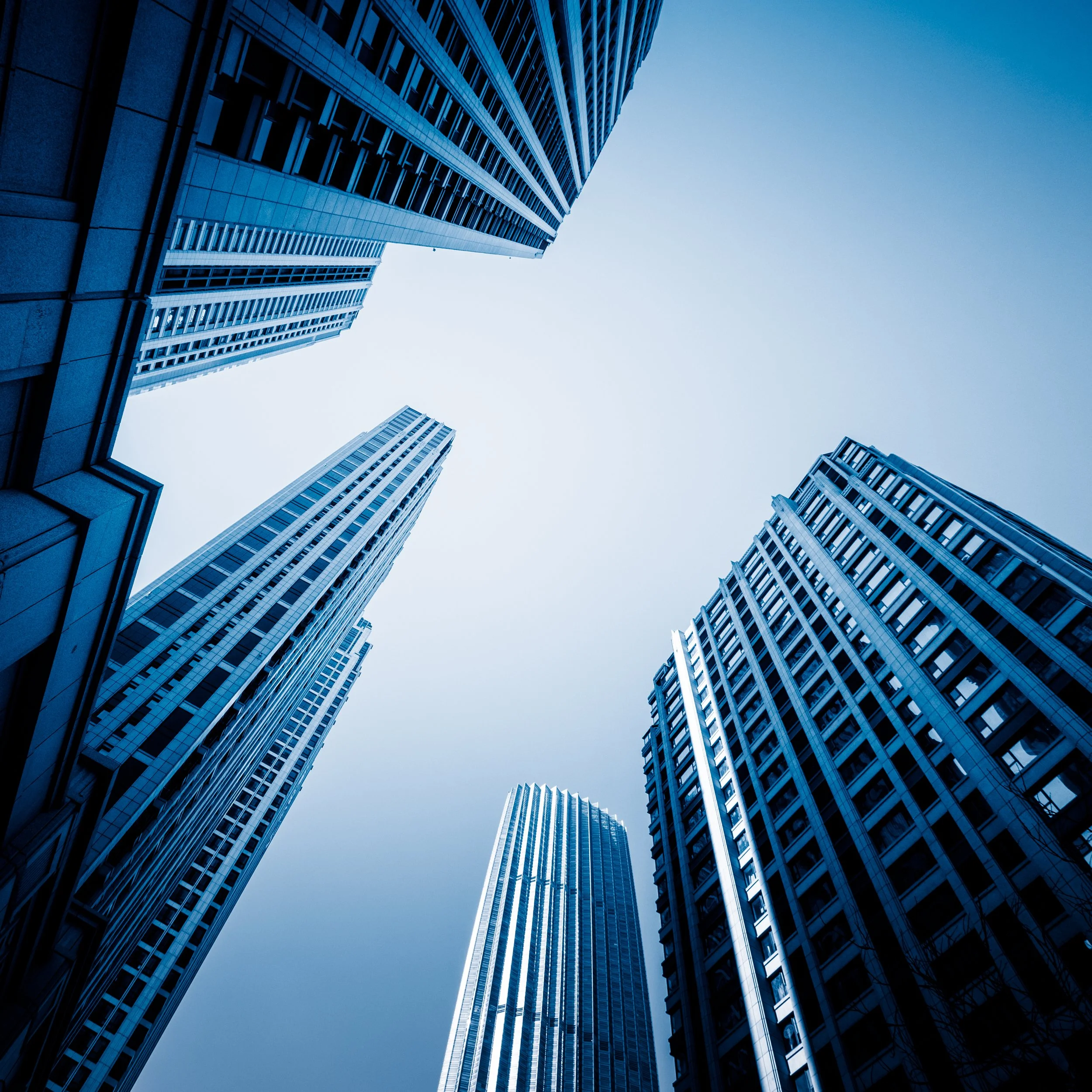 Looking up at several tall skyscrapers in a city, reaching towards a clear, blue sky.