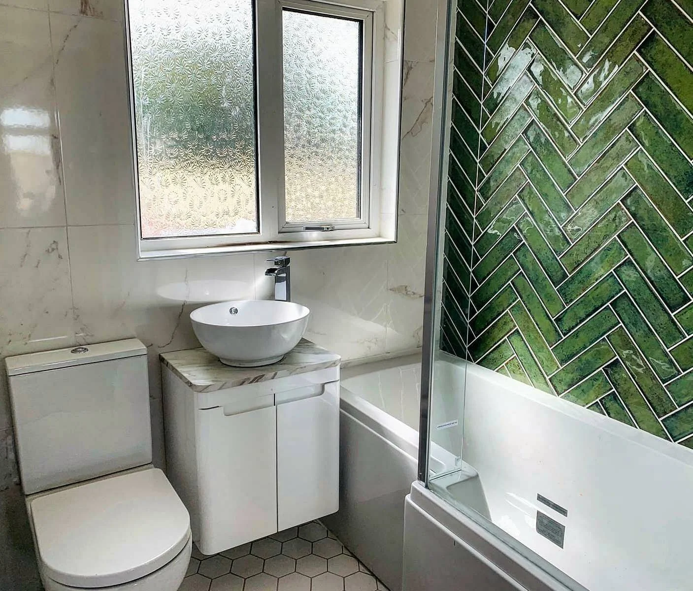 Bathroom with a window, toilet, small sink on a cabinet, bathtub, and green herringbone-patterned tiled wall.