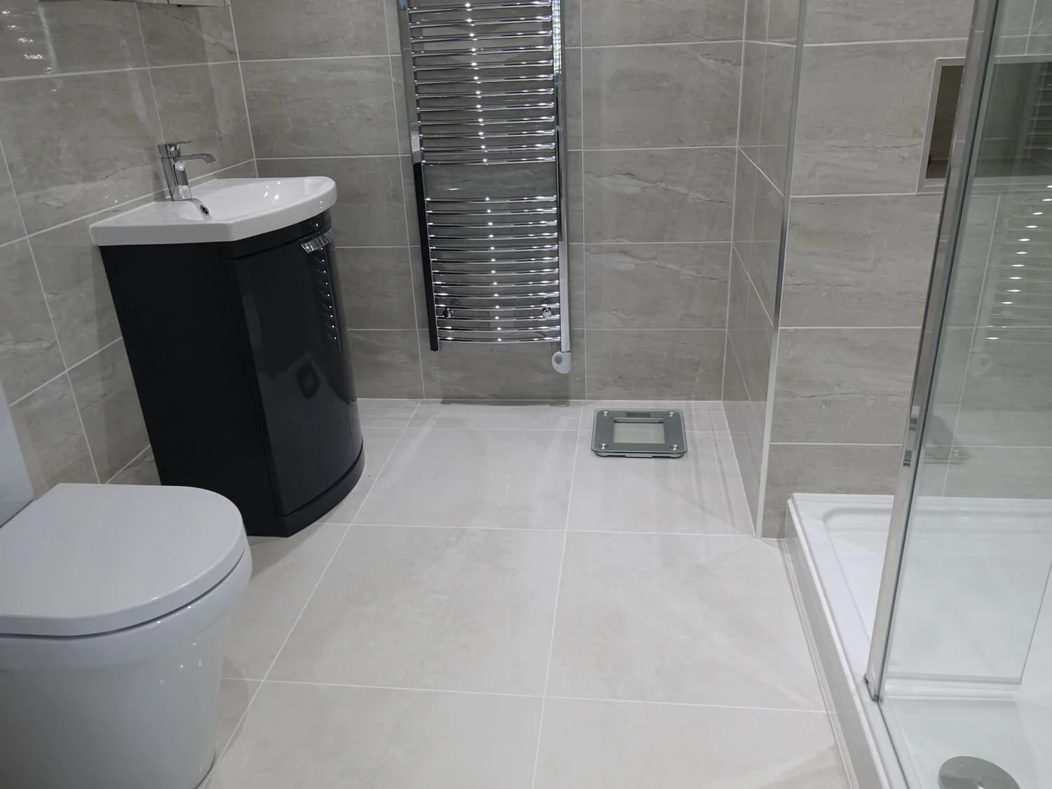 Modern bathroom with gray tiled walls and floors, featuring a white toilet, a black storage cabinet with a sink, a chrome towel radiator, a bathroom scale, and a glass shower enclosure.