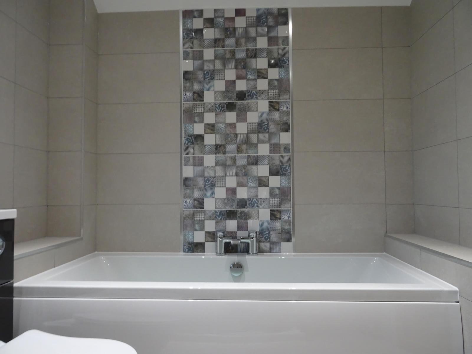 Bathroom with a bathtub, beige tiled walls, and a vertical strip of decorative tiles with various patterns in shades of gray, black, and white.