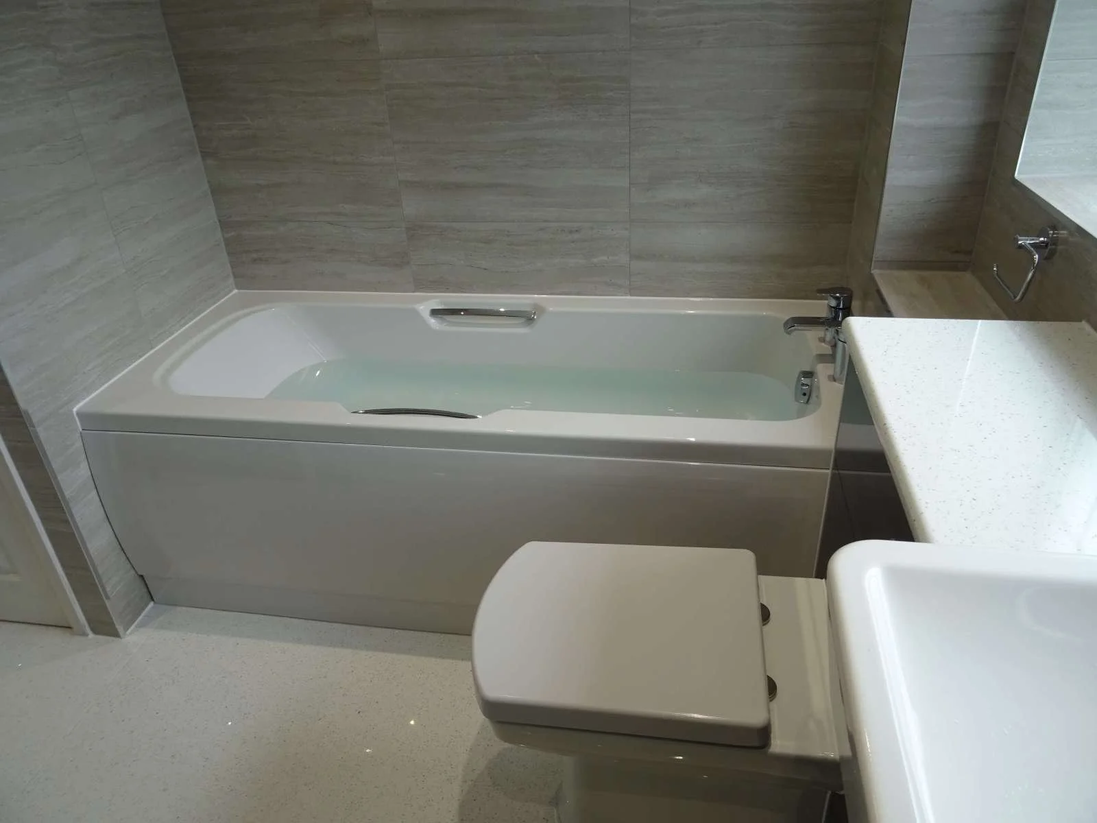 Modern bathroom with a bathtub, a toilet, and a sink. The bathtub has a handlebar and is installed against a tiled wall. The toilet is positioned next to the bathtub, and a white countertop with a faucet is visible.