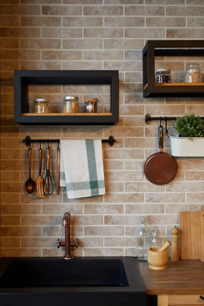 Kitchen with exposed brick wall, black shelves with jars, towel rack with hanging utensils and towel, copper sink, wooden countertop with glass bottles, mortar and pestle, and cutting board.