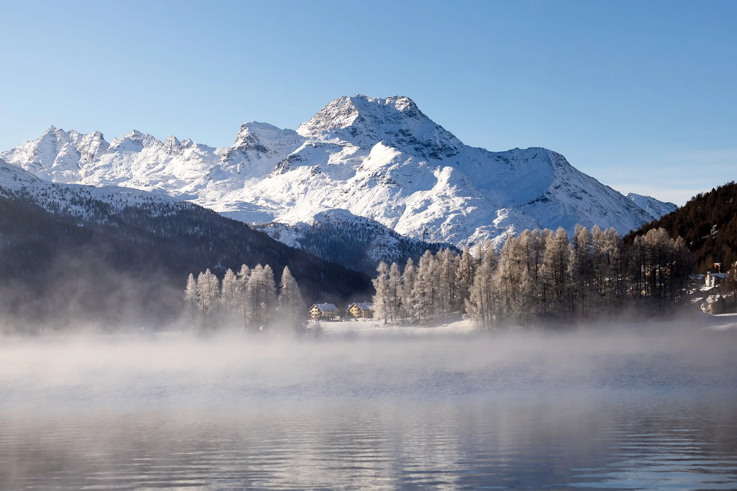 Schneebedeckte Berge im Hintergrund mit einem kleinen Dorf und Bäumen im Vordergrund, umgeben von Nebel über einem ruhigen See.