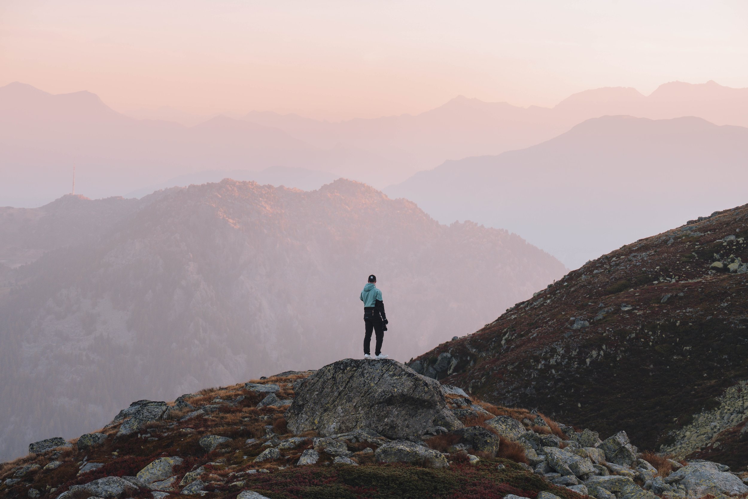 Ein einzelner Wanderer steht auf einem felsigen Hügel und blickt auf eine nebelverhangene Berglandschaft während des Sonnenaufgangs oder Sonnenuntergangs.