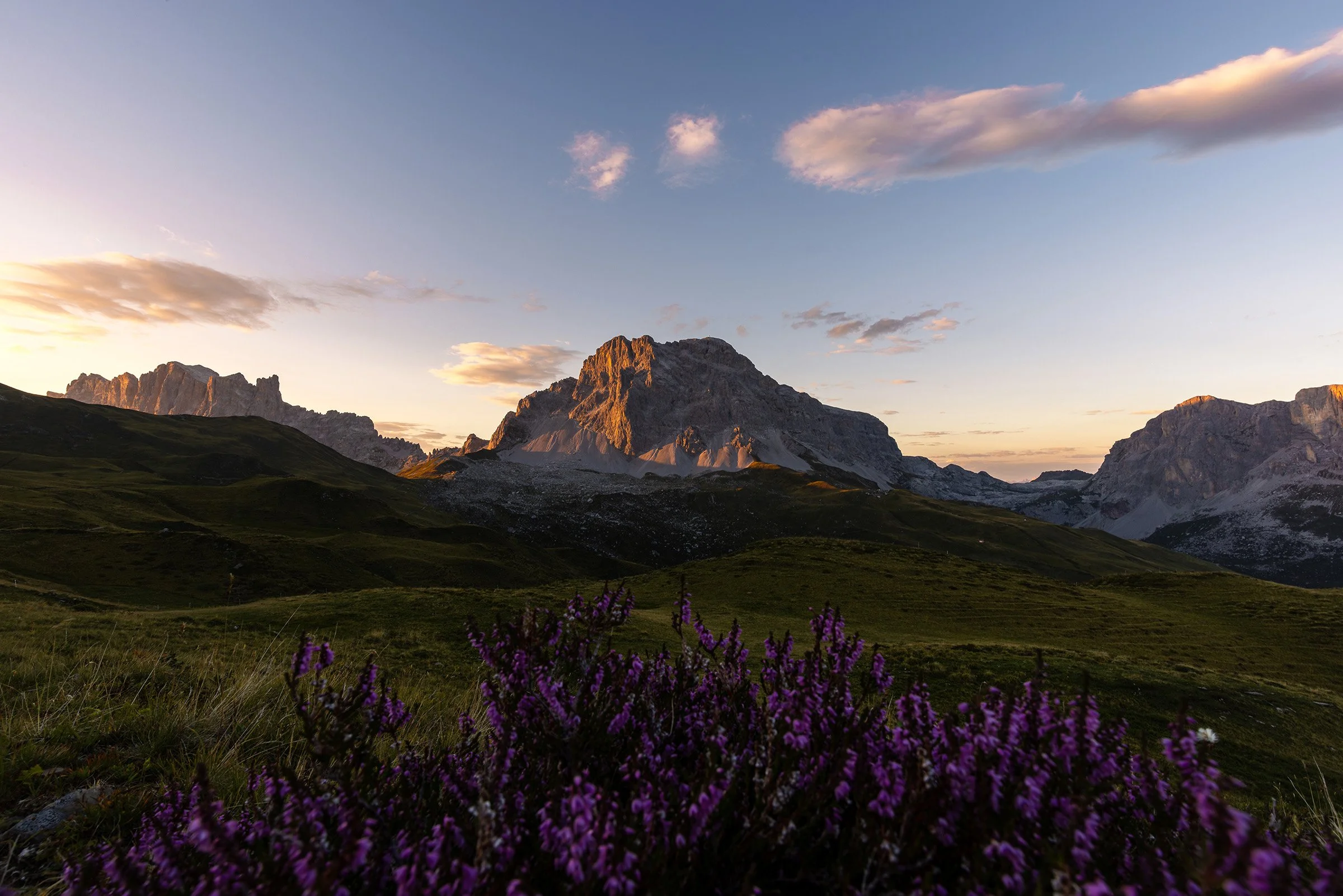 Eine bergige Landschaft bei Sonnenuntergang mit grünen Wiesen, Felsen, Bergen und einem blauen Himmel mit Wolken.