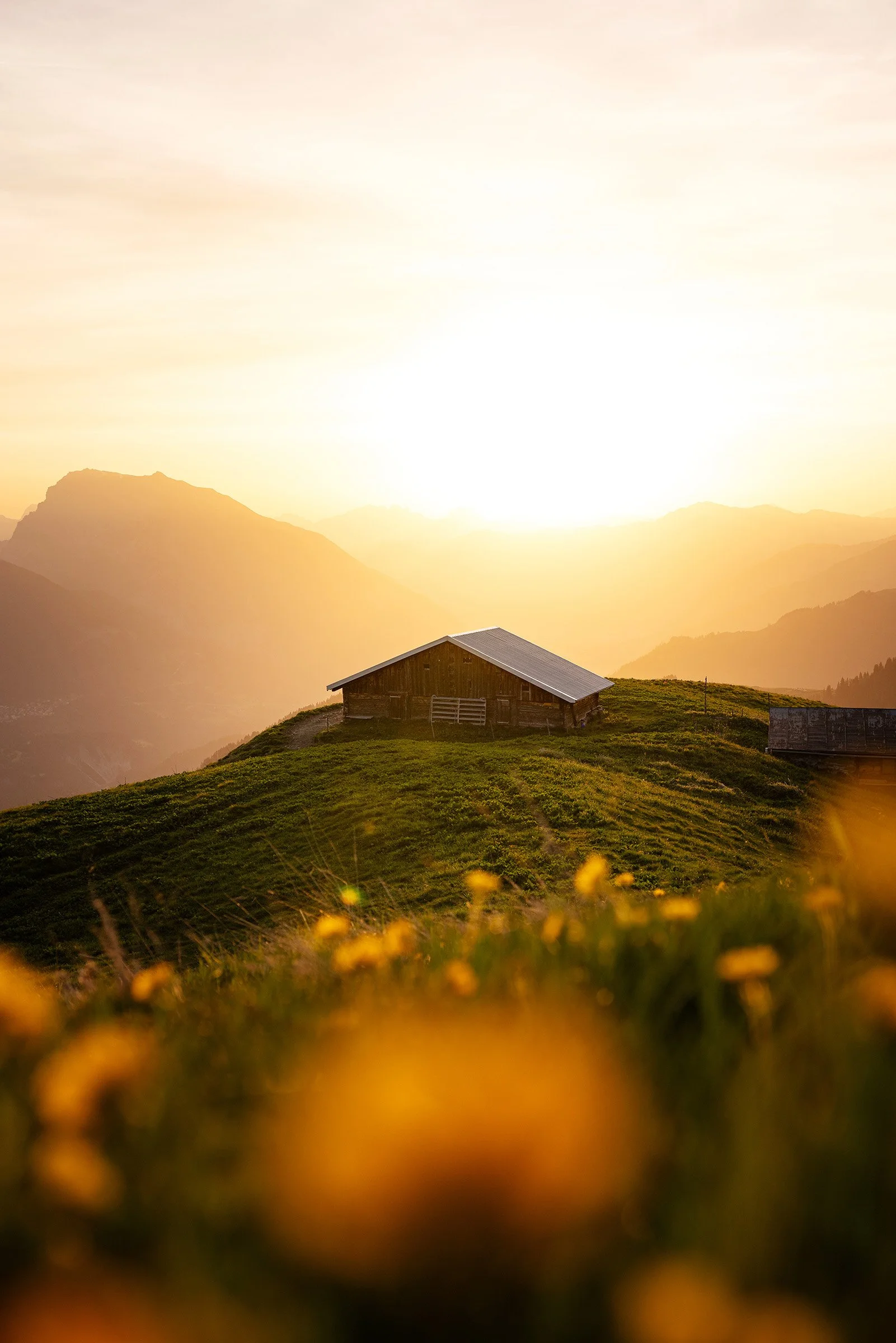 Sonnenuntergang über Bergen mit einem Bauernhaus im Vordergrund auf einer grünen Wiese.