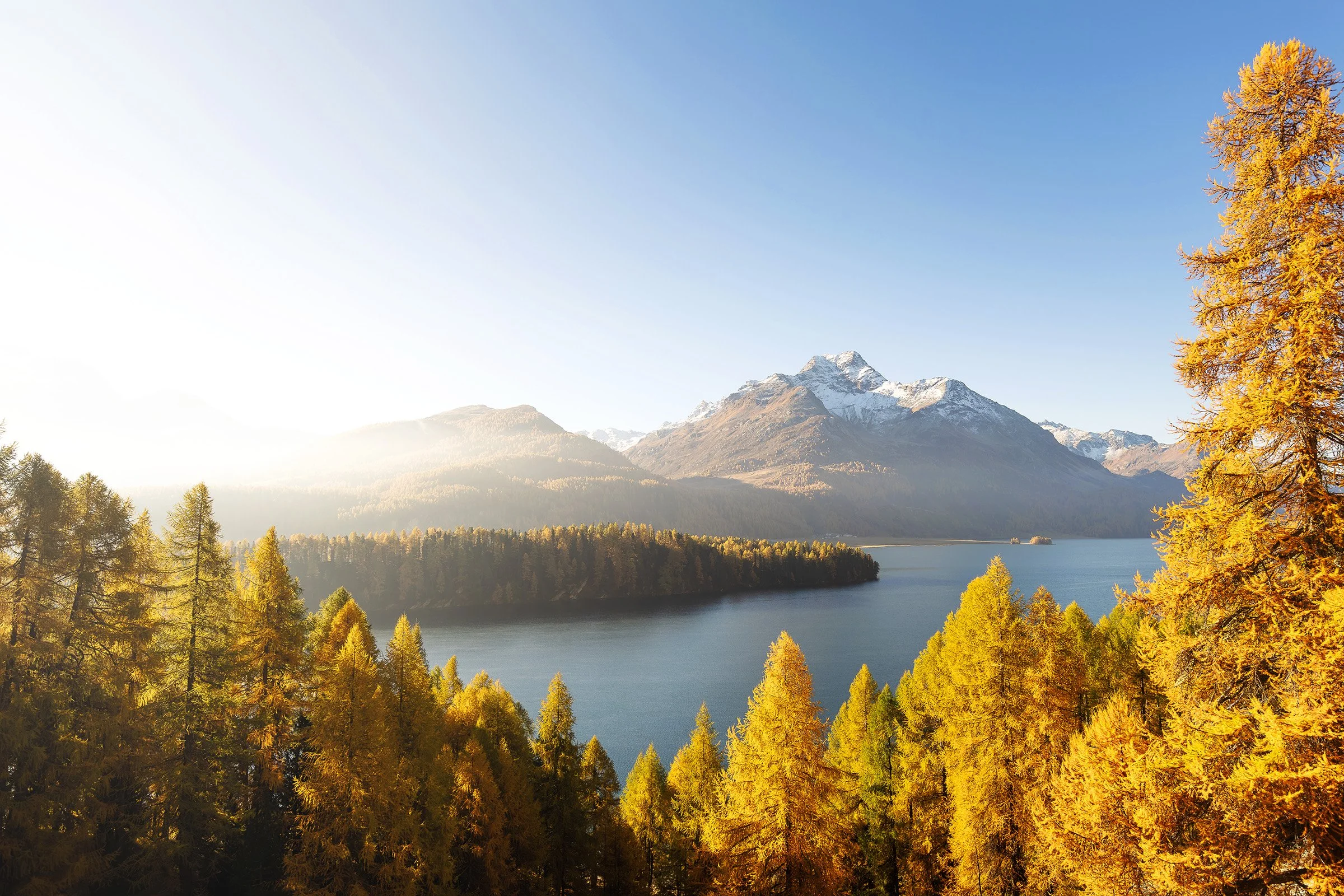 Ein See im Tal, umgeben von einer Kette von Bergen mit schneebedeckten Gipfeln. Im Vordergrund befinden sich zahlreiche Bäume mit goldgelben Blättern, die auf den Herbst hinweisen.