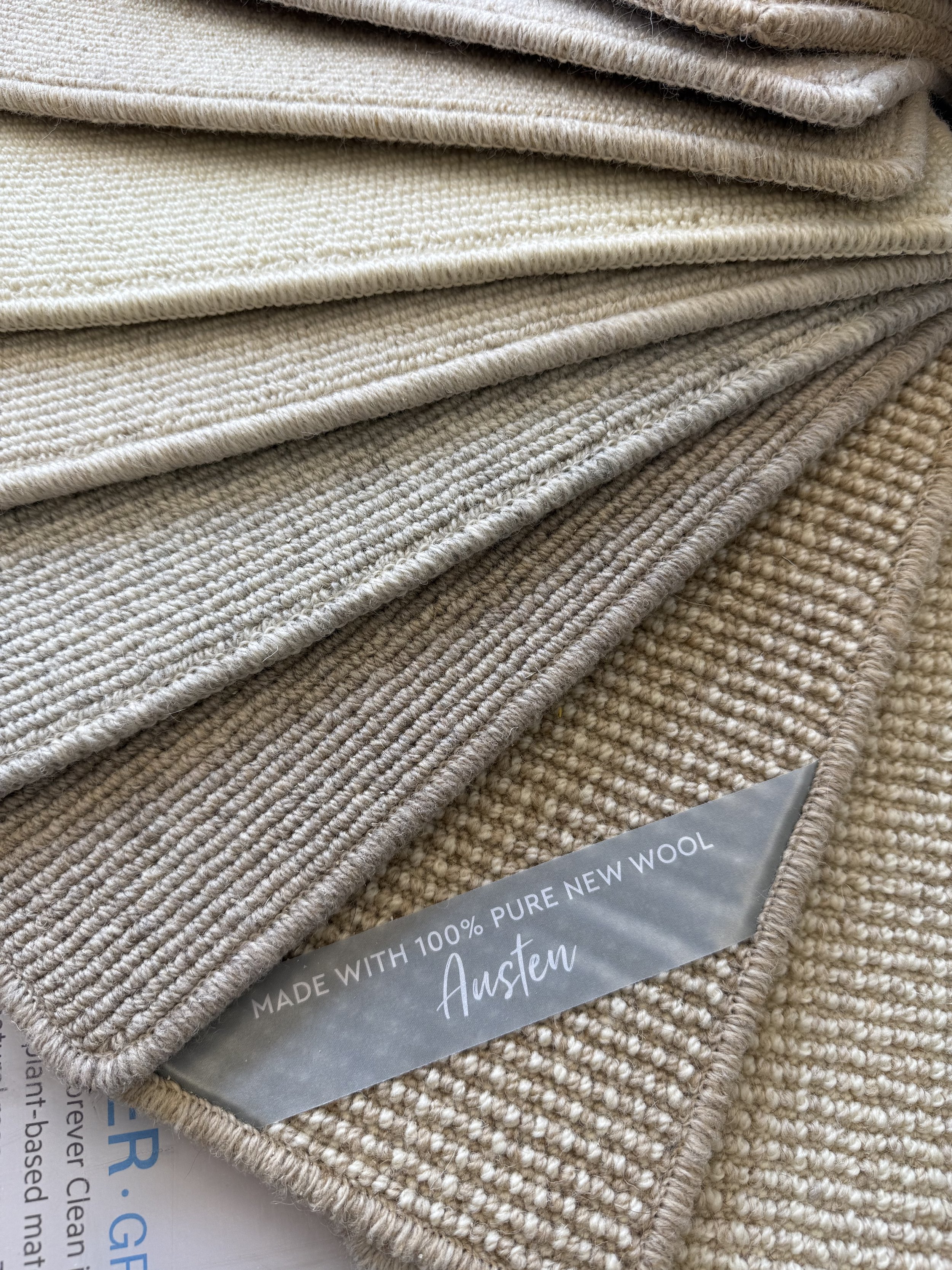 Cormar Carpet - Stacked swatches of beige wool fabric showing different textures and weaves, with a label indicating they are made from 100% pure new wool and branded 'Austen.'