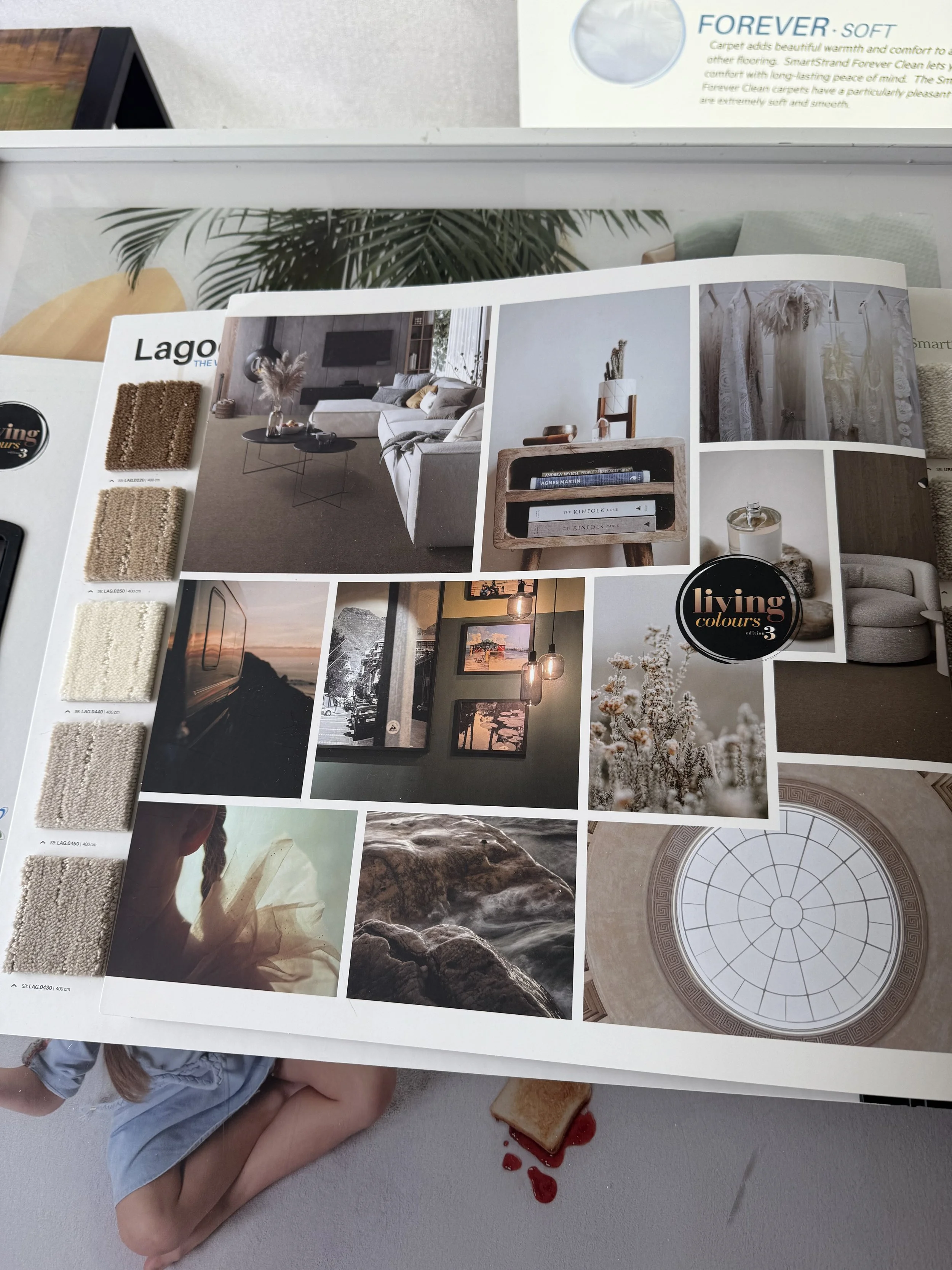 A home decor catalog showing interior design images, fabric swatches, and a "living colours" logo, overlaid on a table with a child's leg, a slice of toast with jam, and a child in the background.