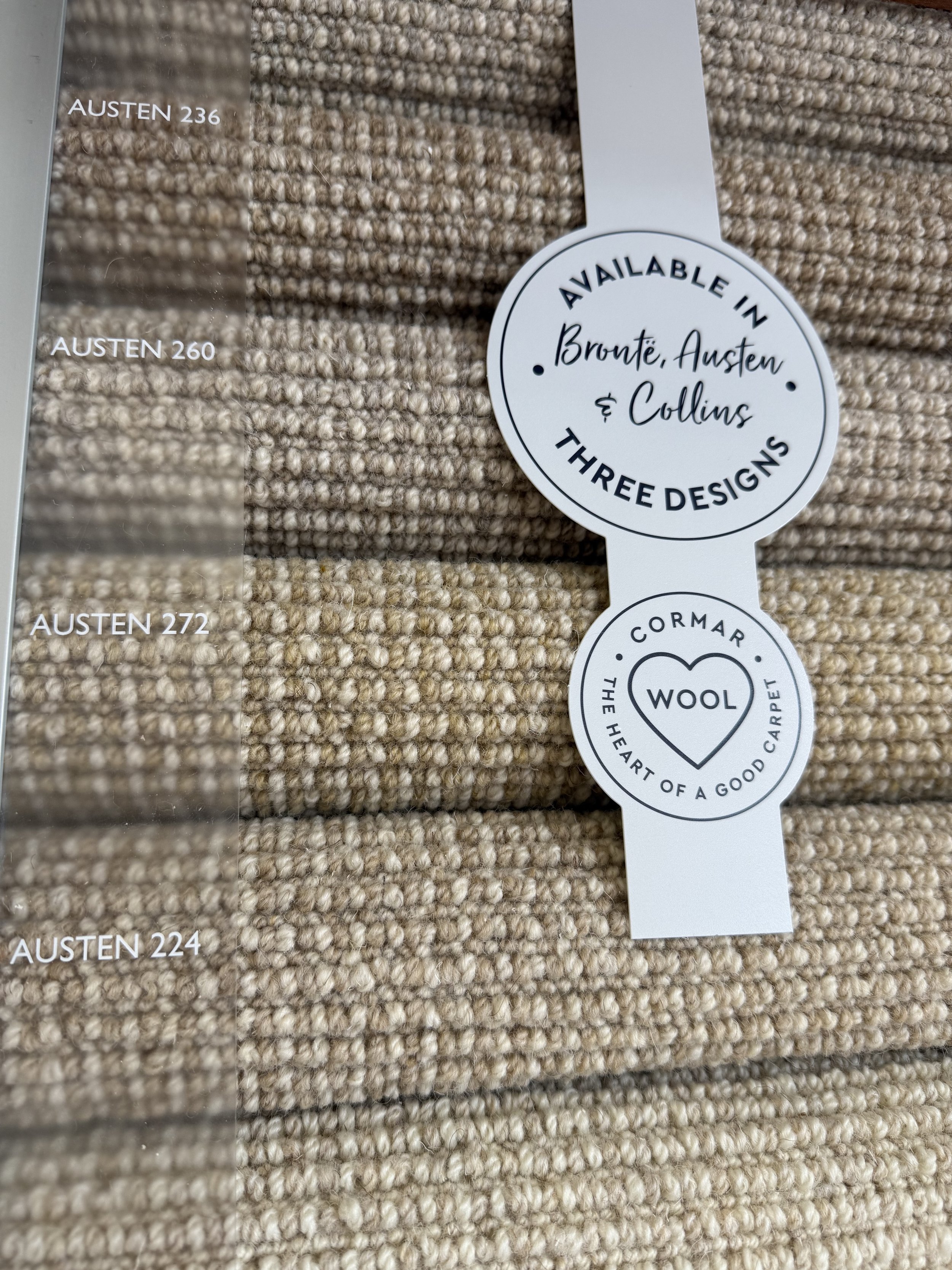 Close-up of a carpet sample display with four beige woven carpet swatches labeled AUSTEN 236, AUSTEN 260, AUSTEN 272, and AUSTEN 224. A circular tag states the designs are available in Bronte, Austen, and Collins, and features the Cormar Wool logo an