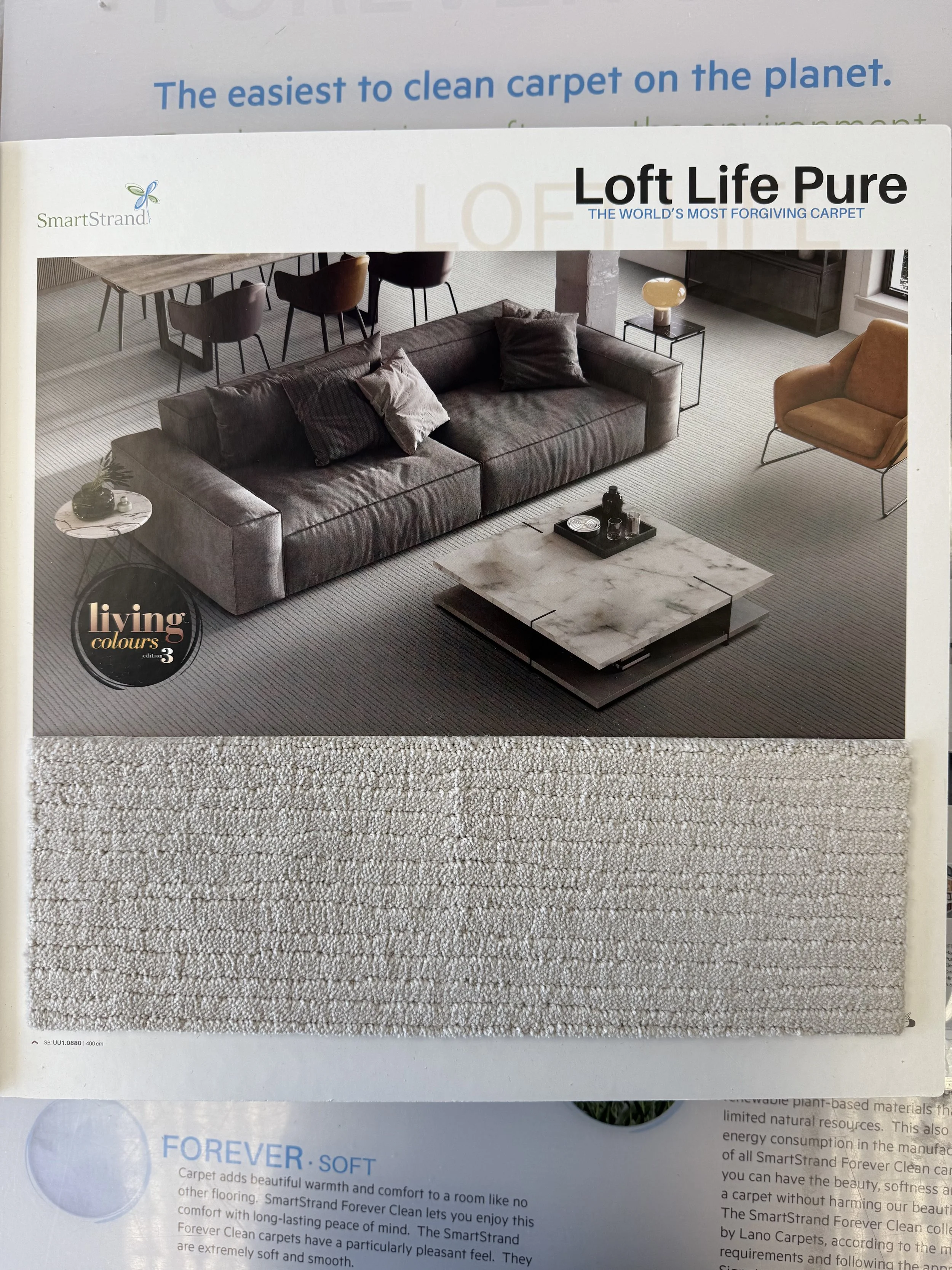 SmartStrand Carpet - A carpet sample and a living room photo on a magazine page. The room has a dark sofa, a round marble coffee table, a beige ottoman, and a dining area. The carpet sample shows a textured, neutral-colored, plush carpet.
