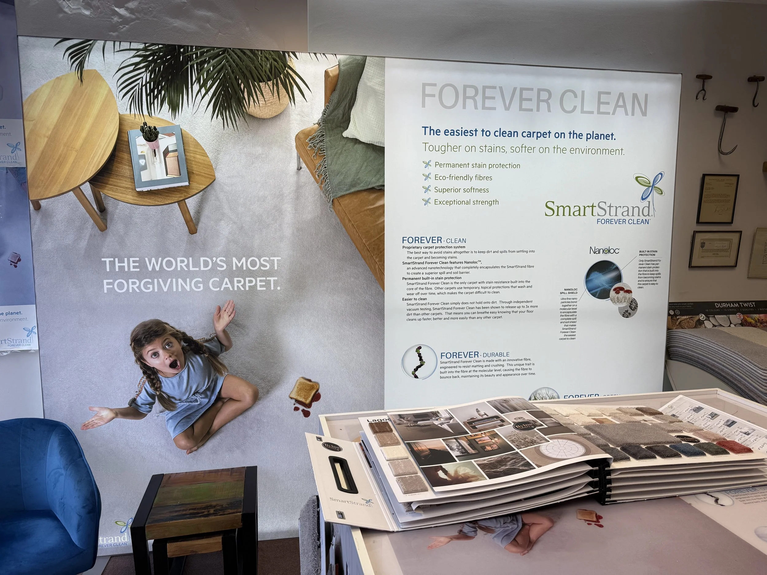 A young girl sitting on a light-colored carpet in an office or showroom, looking up with a surprised expression. There is a slice of toast with jelly on the carpet, with jelly spilled next to it. The room contains two small wooden tables, a blue chair, and a large poster advertising carpet cleaning products and services. The poster has text about a Forever clean and durable carpet system, with a logo labeled SmartStrand.