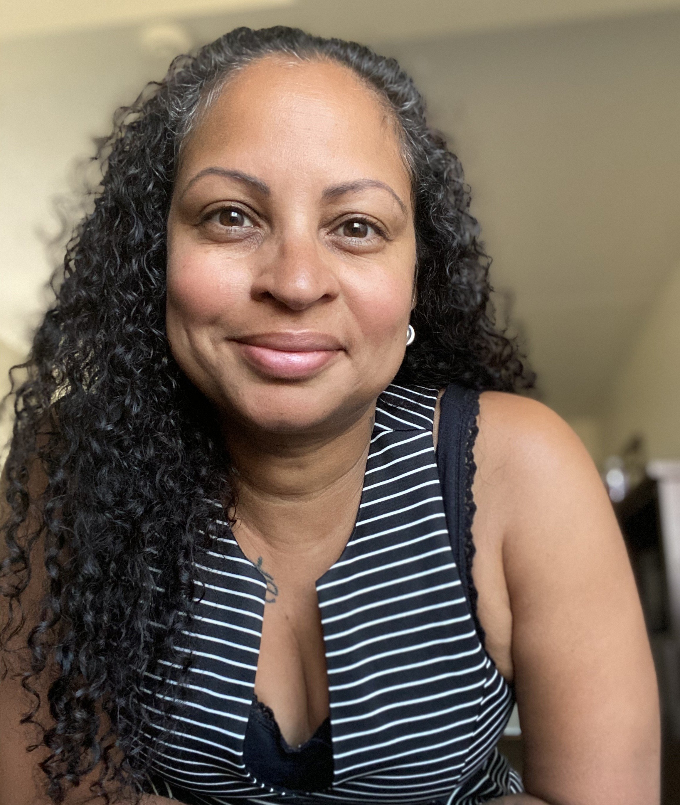 A woman with curly black hair and light brown skin, smiling, wearing a black and white striped sleeveless top, sitting indoors with a beige wall in the background.