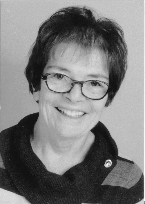 Cheryl Cavell - A woman with short hair, glasses, and a dark patterned sweater smiling in a black and white portrait.