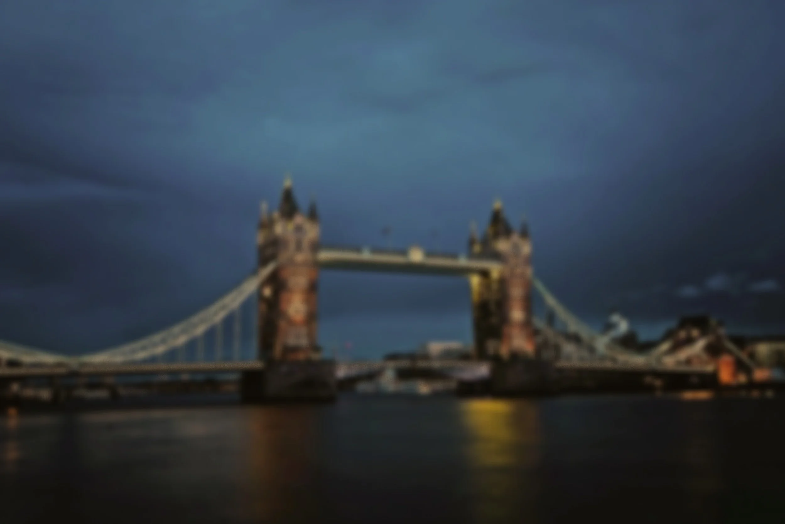 A blurry nighttime photo of Tower Bridge in London with dark clouds in the sky and reflections on the river below.