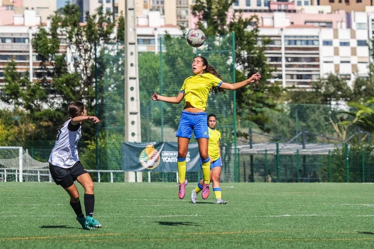Girls’ football match action with a player jumping to head the ball during the Lisbon Football Youth Cup 2025.