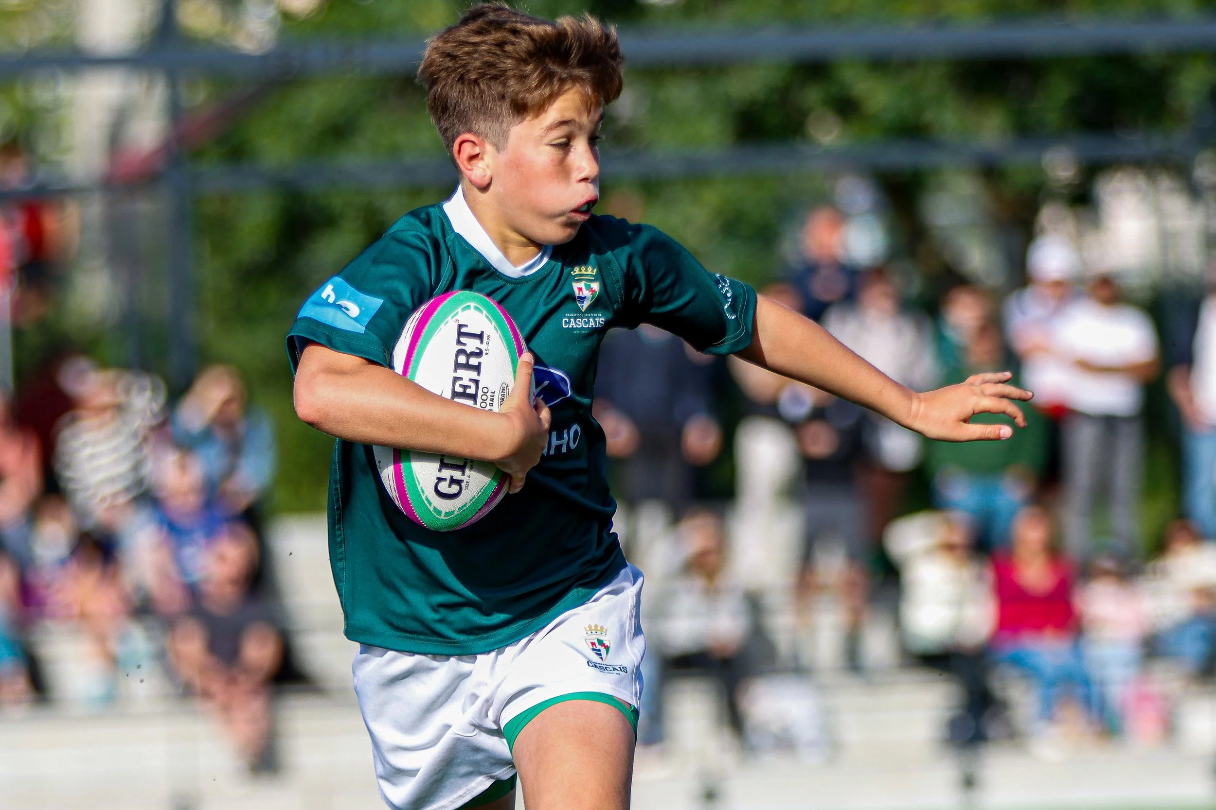 A young boy in a green sports uniform running with a rugby ball while a crowd watches on the sidelines.
