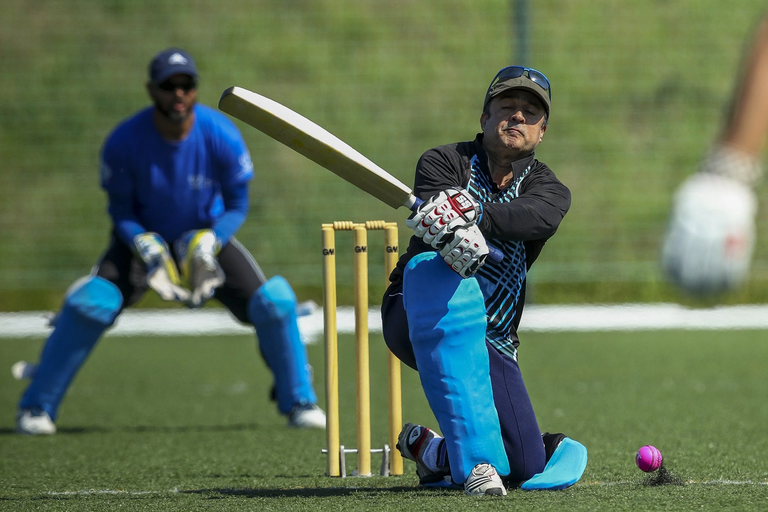 Cricketer batting with a pink ball during practice or match, with wicketkeeper in background on grassy field.